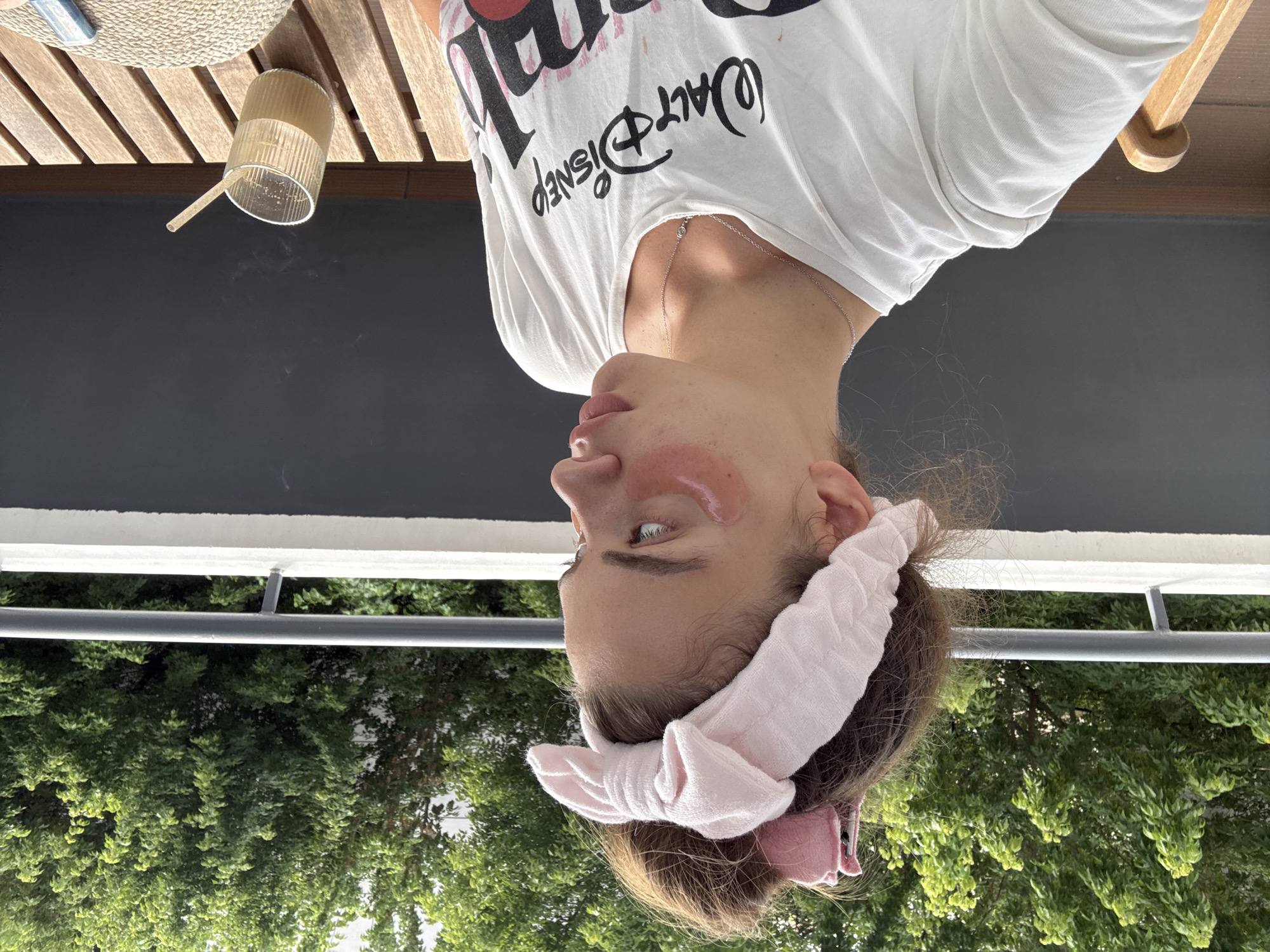 A young woman with brown hair tied up and a pink headband with a bow is sitting on a balcony in Plovdiv, Bulgaria. She is wearing a white t-shirt with a Walt Disney graphic. She has pink under-eye patches and is looking to the right. A glass with a drink and a straw is on a wooden table next to her. Lush green trees and a balcony railing are visible in the background.