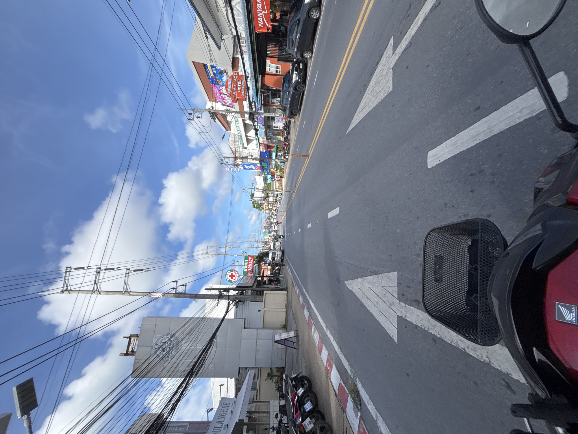 The image captures a street view in Chalong, Thailand, likely on Soi Nanai, a bustling road known for its shops and hotels. The perspective is from the rider of a scooter, with the handlebars and a black basket in the foreground. The road is paved with asphalt and marked with white lane lines and an upward-pointing arrow. On the left side of the road, a sidewalk is lined with parked motorcycles and businesses, including a "BOULANGERIE" establishment. Further down the street, a red and white striped curb is visible.
On the right side of the road, buildings house various businesses and a hotel. Signage indicates "JULA PLACE HOTEL" and "NANYANG." Traffic is present, with cars and motorbikes visible on the road. Electrical poles and wires crisscross the sky, a common sight in Thailand. The sky is a clear blue, punctuated by fluffy white clouds, indicating fair weather. The scene depicts a typical day in a commercial area of Chalong, characterized by its vibrant street life, diverse businesses, and the ever-present network of electrical infrastructure.