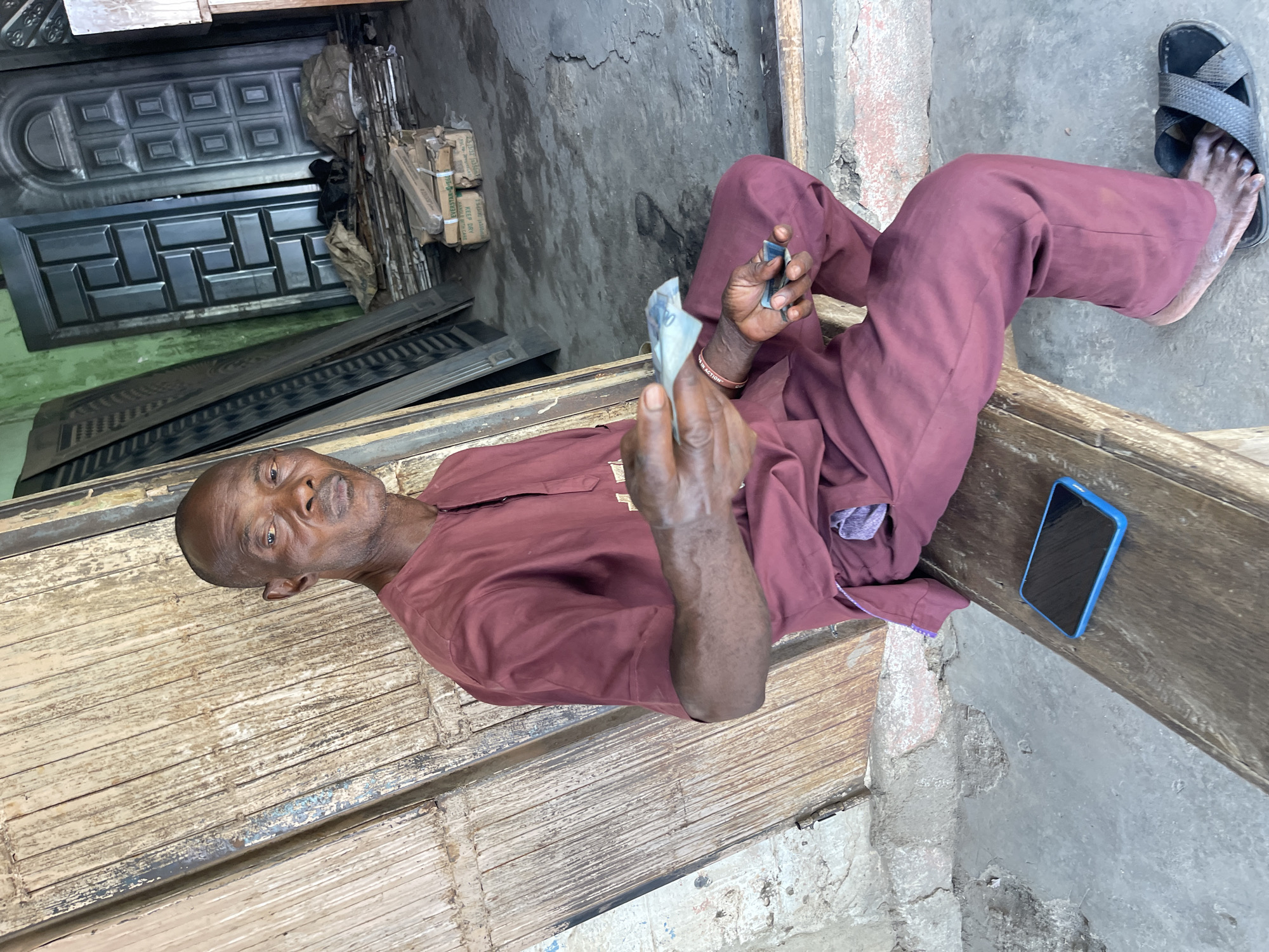 A middle-aged Black man sits cross-legged on a weathered, rough-hewn wooden structure outside a building in Muchiya, Nigeria. He wears a maroon tunic and matching pants. He holds paper money in one hand and a small, rectangular object in the other. Next to him on the structure lies a blue-cased smartphone. His bare foot wearing a black sandal can be seen on the upper right of the frame. Behind him is a gray concrete wall and a stack of metal doors.