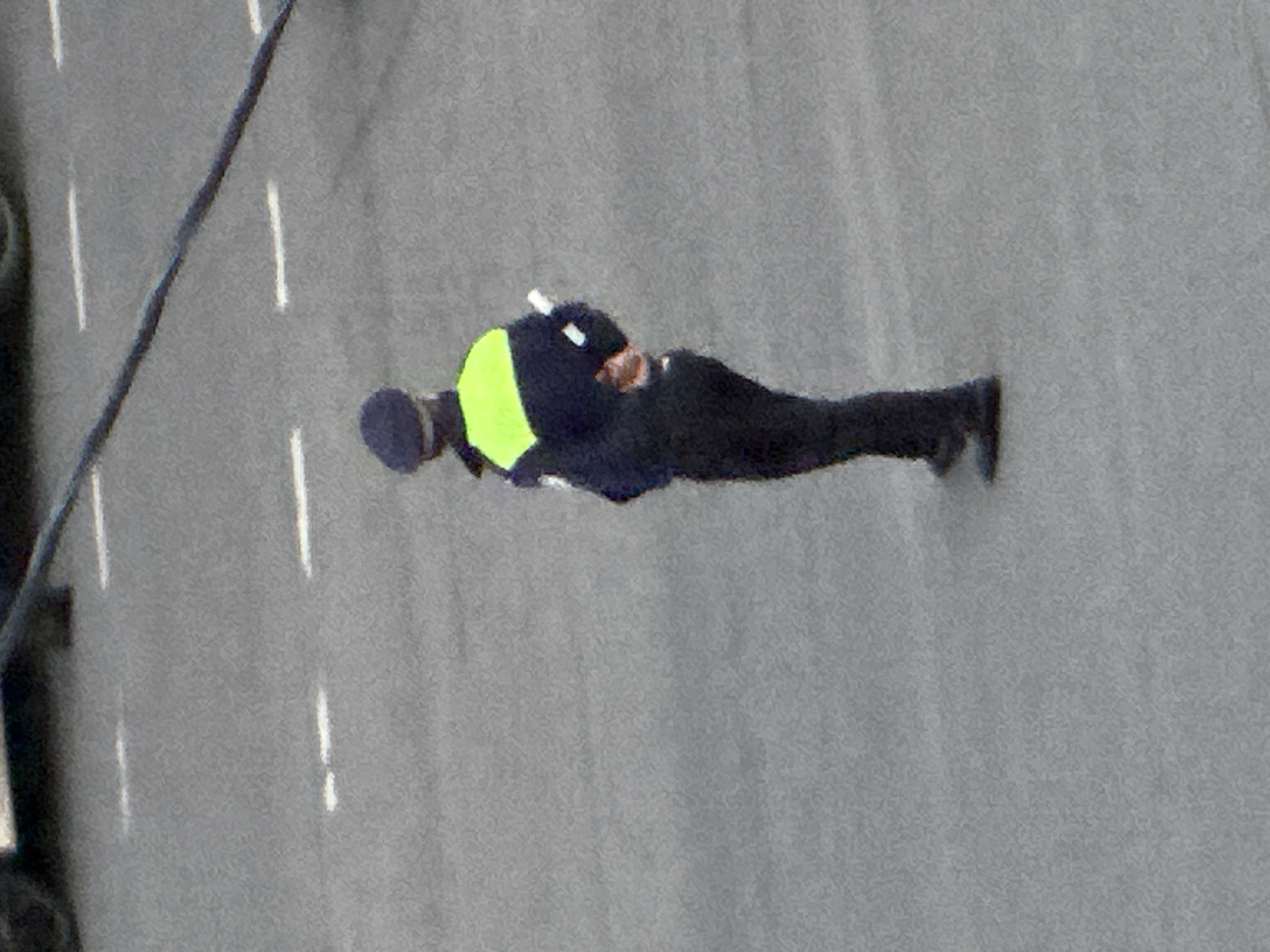 A person in a dark blue uniform with a bright green reflective band across the chest and shoulders, wearing a dark blue cap, stands on an asphalt surface. The person's back is to the camera, and they appear to be holding a white baton or stick in their right hand. The background shows a gray asphalt surface with faint white line markings. The overall scene is outdoors, with a slight grainy quality to the image. There is no specific indication of the city of Podolsk, Russia in the image itself.