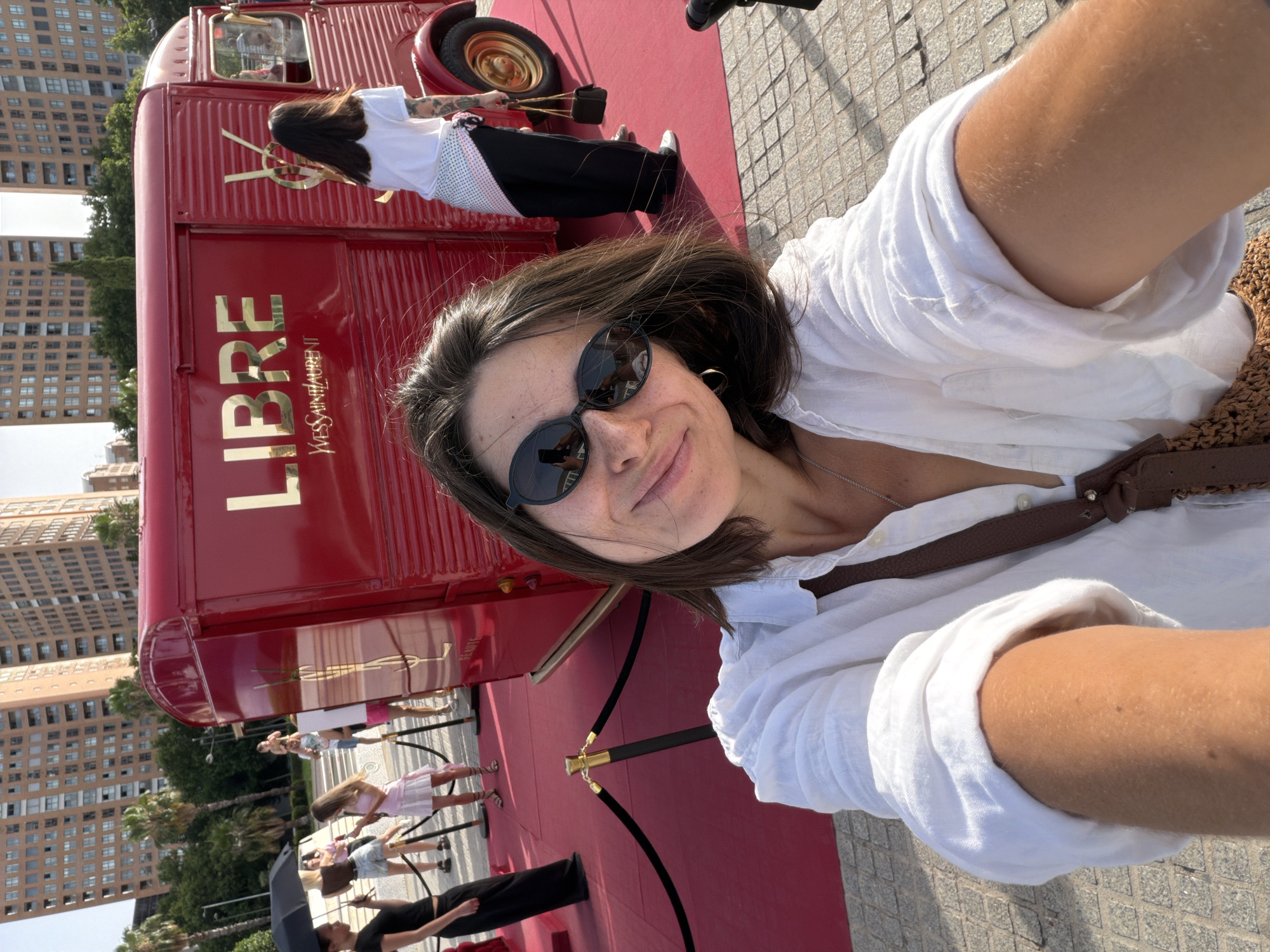 A woman with dark hair and sunglasses is taking a selfie outdoors in front of a red truck with gold lettering that says "LIBRE". She is wearing a white shirt and a brown bag is slung across her shoulder. In the background, there are people walking on a red carpet, palm trees, and tall buildings. The truck appears to be a promotional vehicle for Yves Saint Laurent.