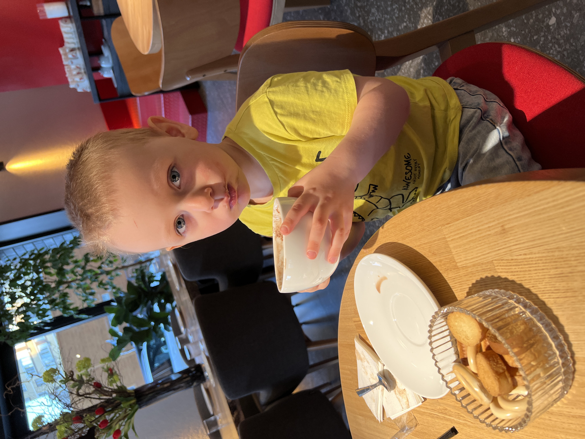 A young boy sits at a light wooden table in a cafe, holding a white mug with remnants of chocolate or coffee around his mouth. He wears a yellow T-shirt and light blue jeans. A white saucer with a spoon and napkin sits next to the mug, along with a glass bowl filled with various cookies and breadsticks. The cafe has other wooden chairs and red seat cushions. A window reveals a view of a building and greenery outside, suggesting an urban setting. This likely doesn't take place in Mytishchi, Russia as nothing in the photo confirms this location.