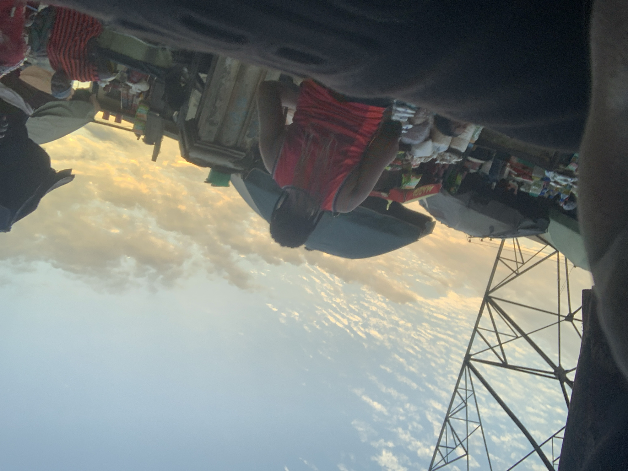 The image captures a market scene in Obosi, Nigeria, during what appears to be sunset, given the warm hues in the sky and the golden light illuminating the clouds. A prominent electrical pylon stands tall on the left side of the frame, its angular structure contrasting with the soft sky. In the foreground, partially obscuring the view, is a dark, out-of-focus mass, possibly the shoulder or arm of a person.
The central focus is a person wearing a bright red tank top, with their back to the camera. They have dark hair that is styled into braids or dreadlocks. This individual is standing amidst market stalls. To the right of the person in the red top, a woman wearing a striped red and black garment is visible, looking towards the camera. Market stalls with various goods are spread throughout the scene, some covered by umbrellas or awnings. Bags of what appear to be produce or commodities are stacked, and there are glimpses of other individuals in the background, engaged in market activities. The overall atmosphere suggests a bustling commercial environment at the end of the day.