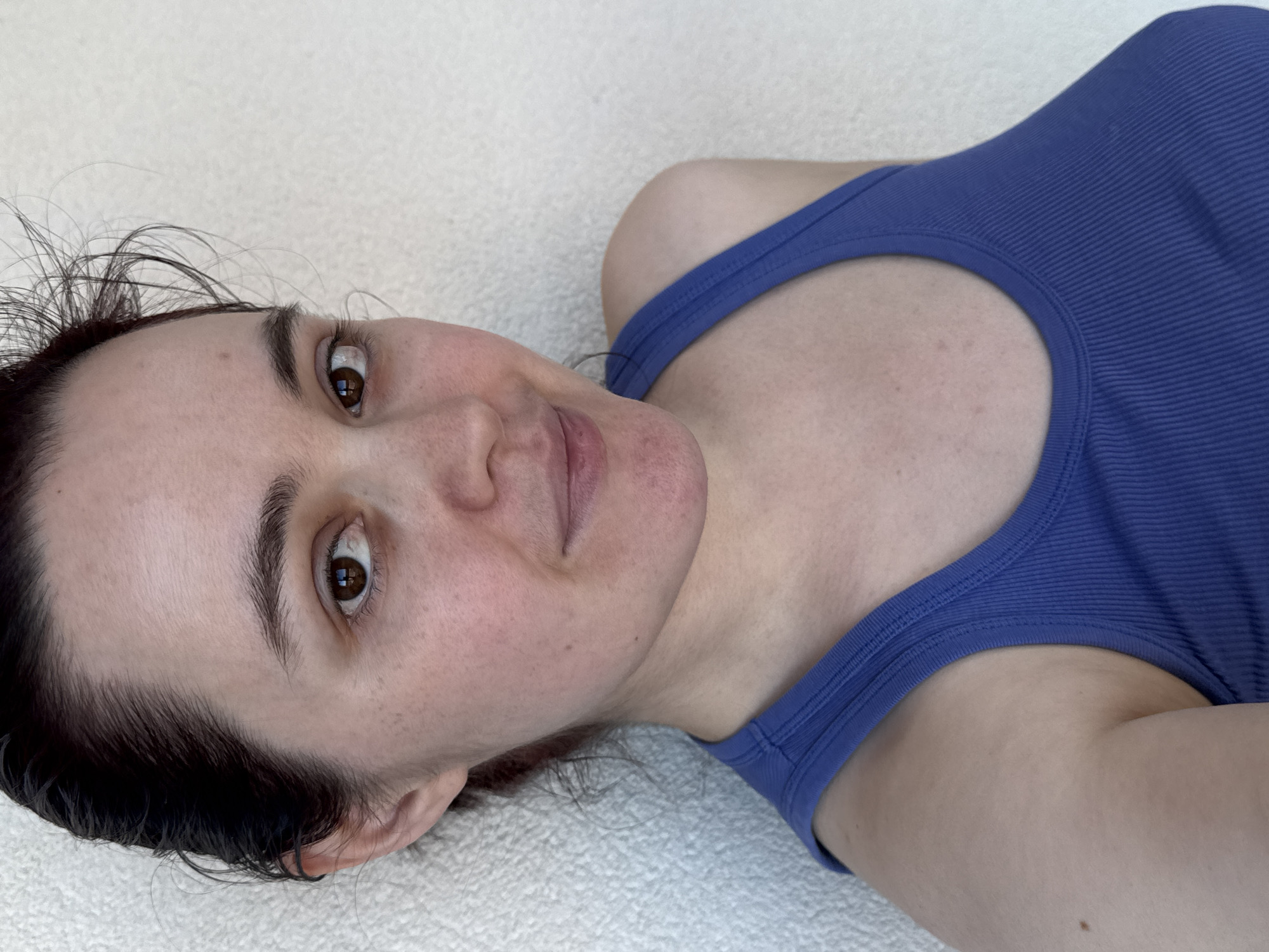 A woman with dark hair and brown eyes is lying on her back on a light-colored textured surface. She is wearing a blue tank top. The setting is unknown and there is no way to know if the setting is Izhevsk, Russia.