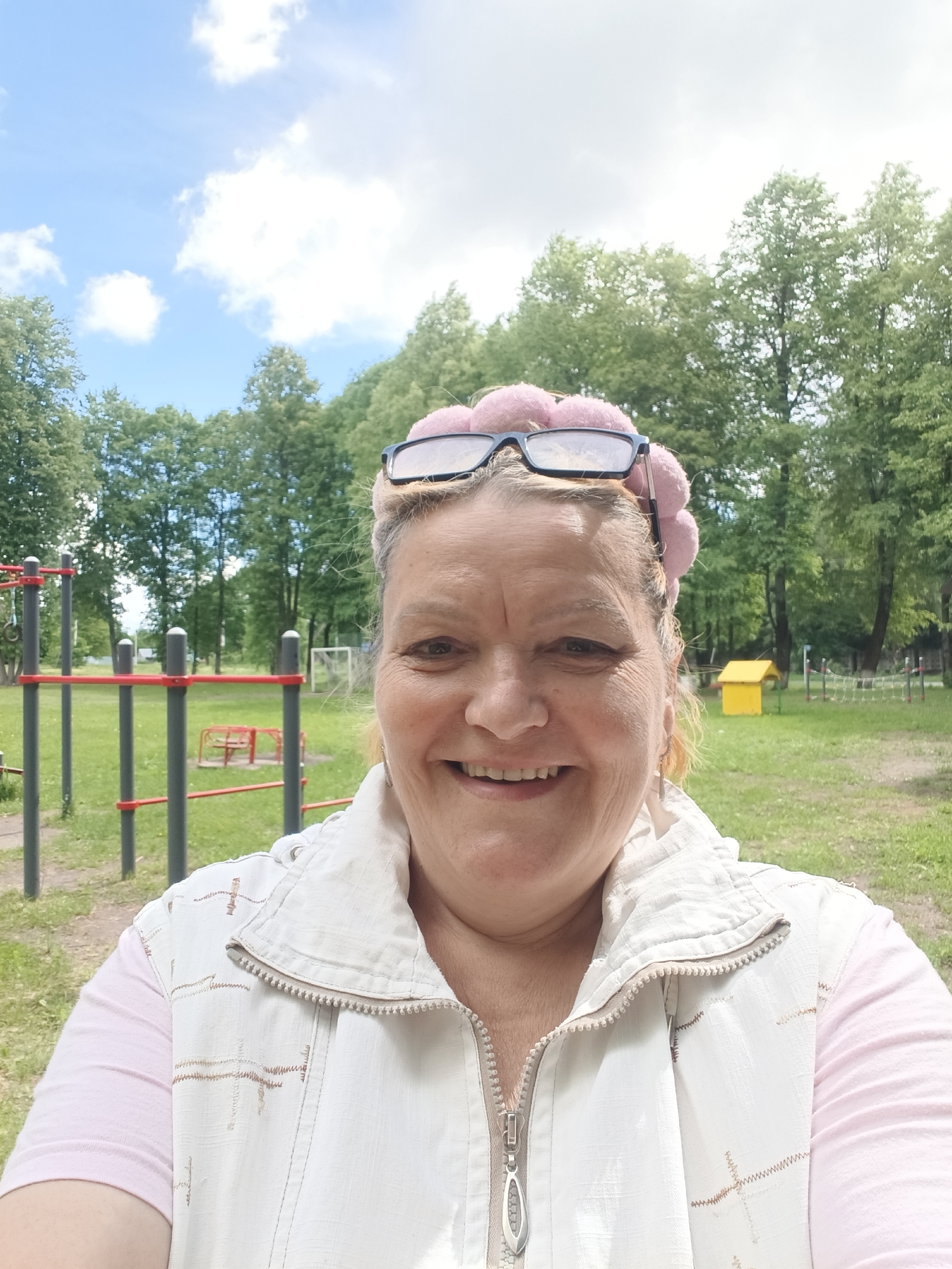 A woman is taking a selfie outdoors in Красный Маяк, Russia. She is smiling and wearing a light pink shirt under a white vest. She has glasses on her head and a decorative pink headband with pom poms. The background shows a playground with a climbing structure with red horizontal bars, a small yellow house-like structure, and green grass. Trees are visible behind the playground, and the sky is blue with some white clouds.