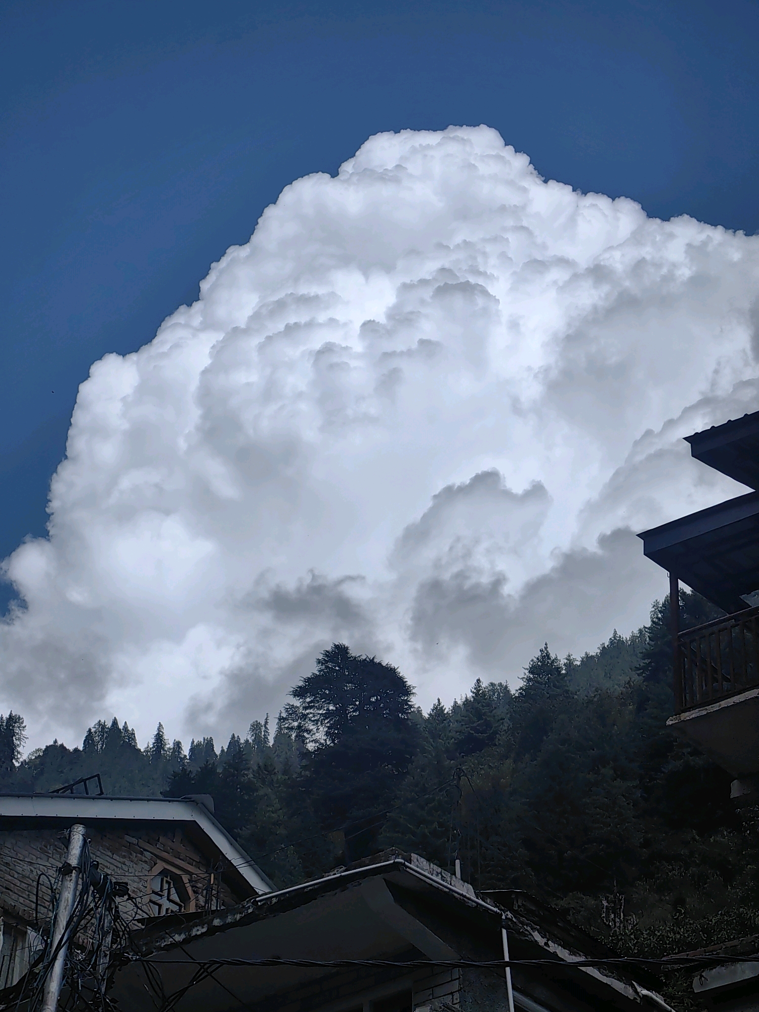 In Vashisht, India, a dramatic sky dominates the scene, filled with a massive, luminous white cloud formation that stretches across the upper portion of the frame. This cloud is textured and appears to be a cumulus cloud, possibly a cumulonimbus, given its size and dramatic appearance. Below the cloud, a dense forest of dark green pine trees covers a hillside, creating a stark contrast to the bright white of the cloud. In the foreground, two buildings with pitched roofs are visible. The building on the left is constructed with brickwork and has visible electrical wires and conduits attached to its facade. The building on the right has a darker roofline and a wooden balcony with railings. The overall impression is one of a mountainous landscape under a dynamic and imposing sky.