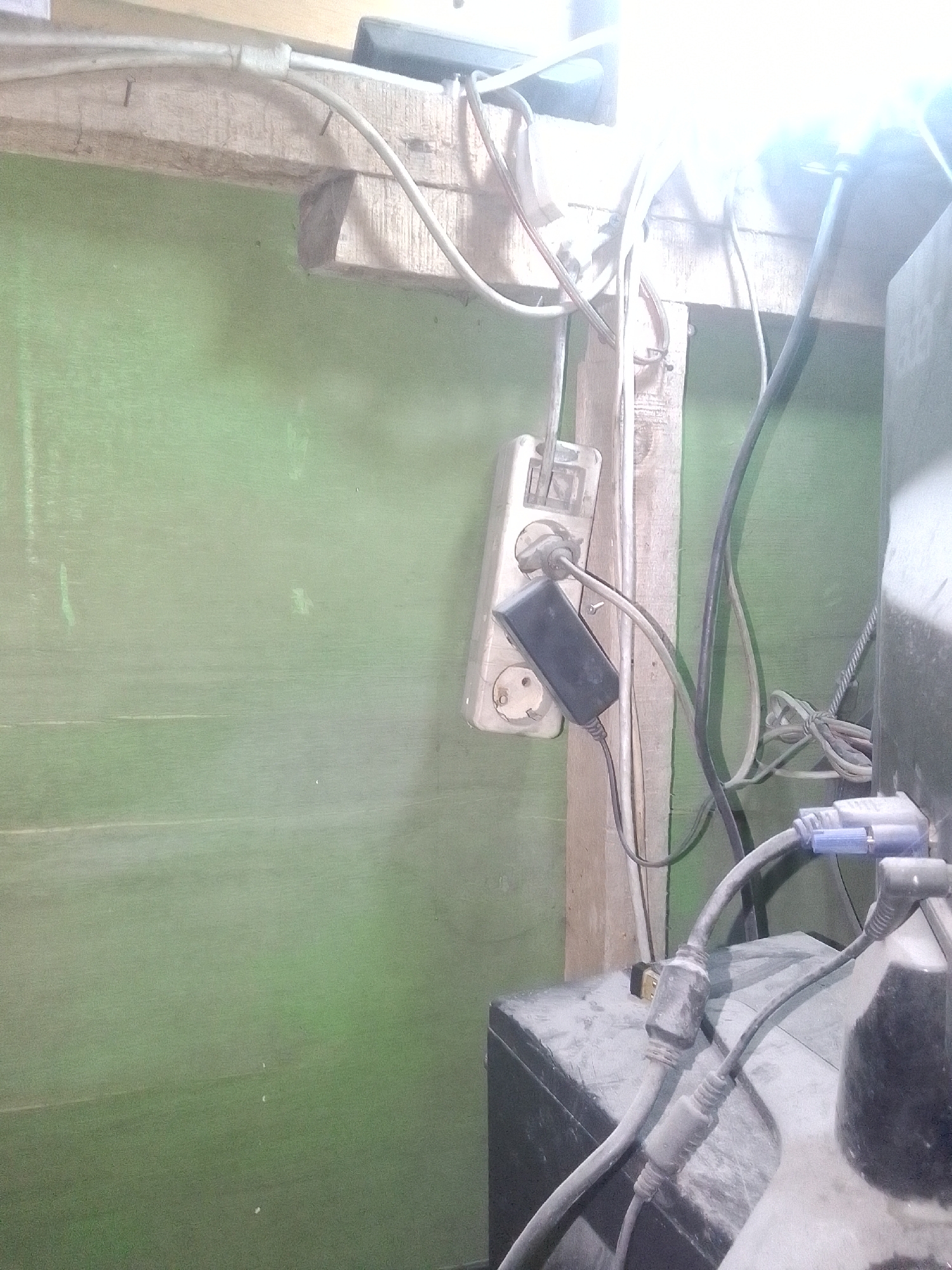 The image depicts a close-up, eye-level view of a cluttered electrical setup. In the foreground, a black electronic device, possibly a computer tower or a large speaker, is covered in a layer of dust. Several cables connect to this device. One prominent cable is gray with a blue connector, likely a VGA cable, plugging into the device. Another gray cable with a black connector, possibly an HDMI or USB cable, is also visible. A thick black cable runs vertically along the right side of the frame.
Behind the electronic device, a white electrical outlet strip is mounted on a wooden beam. A black power adapter is plugged into the outlet strip, with its own cord extending downwards. Various other cables, both white and gray, are seen connected to the outlet strip and running overhead, secured by tape or ties to wooden beams. The background consists of a plain, light green wall.
The scene appears to be an indoor environment, likely a workspace or an area where electronics are housed. The presence of dust suggests the area may not be regularly cleaned or is in a location prone to dust accumulation. There are no people visible in the image. The overall impression is one of a functional, yet somewhat disorganized, collection of electronic equipment and wiring.
The provided location of Gunungmasigit, Indonesia, does not offer specific details relevant to the visual content of this image. The image itself does not contain any landmarks, signage, or environmental cues that would definitively place it in that specific city or region. It is a generic depiction of an indoor electrical setup.