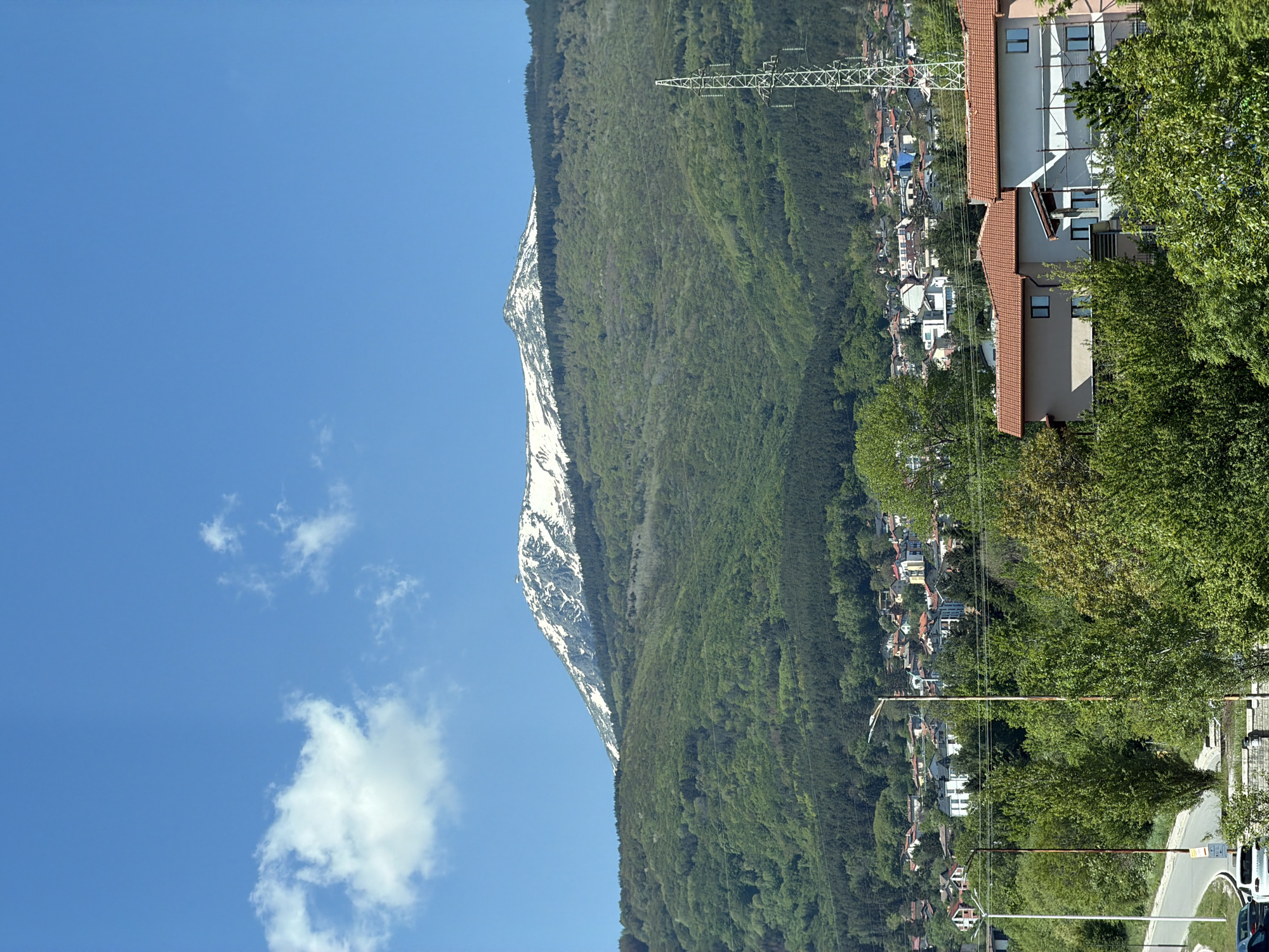 The image depicts a mountainous landscape with a clear blue sky. A peak covered in snow is visible on the left, transitioning into a lush, green mountain slope covered in trees. A town or village is nestled at the base of the mountain, consisting of various buildings with different colored roofs. An electrical tower is situated on the right side of the image. A residential area is seen in the foreground on the right, featuring a large building with a red tile roof and green foliage. The location is likely a mountainous area in the vicinity of the city of Sofia, Bulgaria, with Vitosha Mountain visible in the background.
