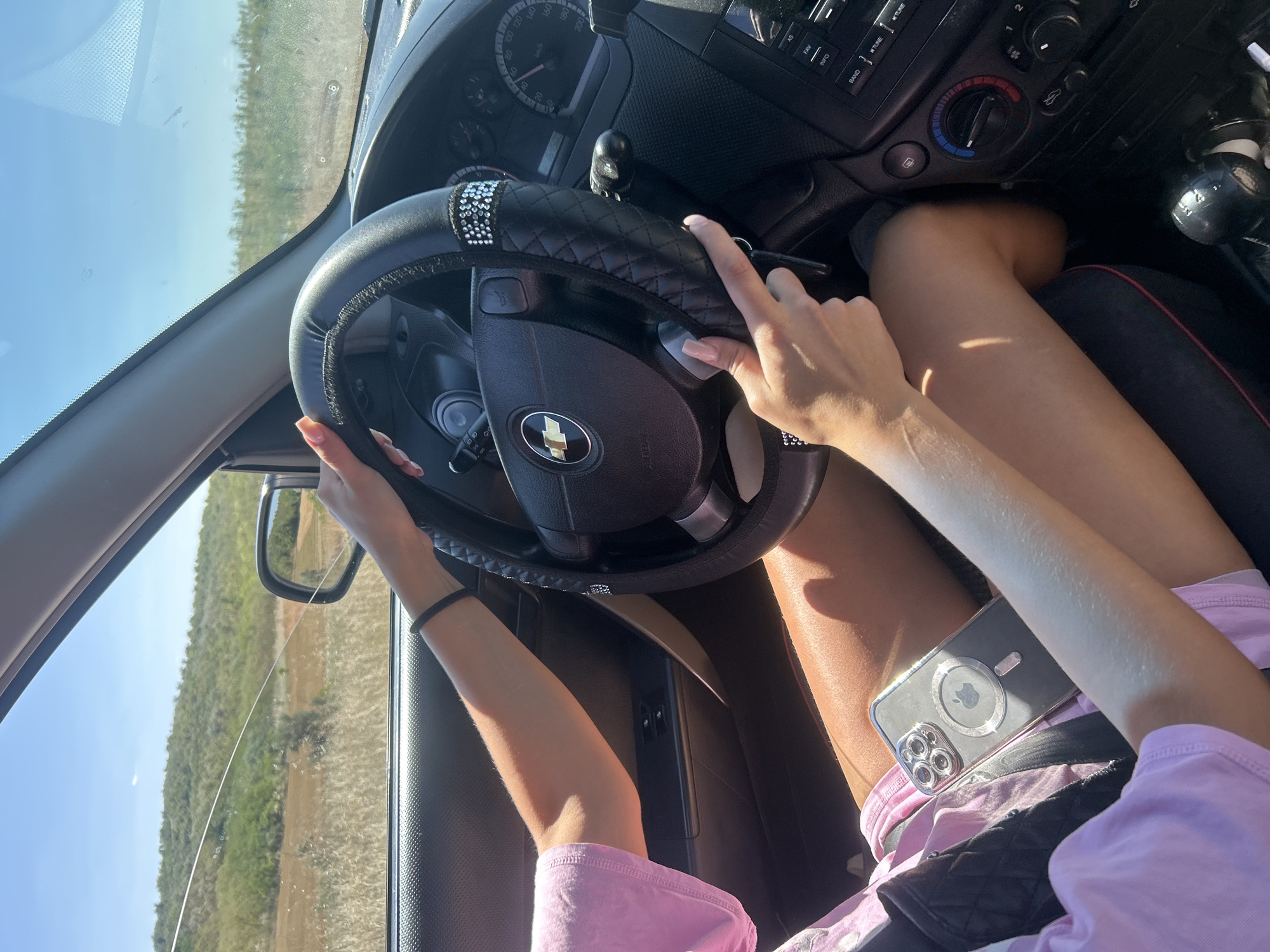 In a car interior, a person with tan skin is driving on a sunny day. They are wearing a pink t-shirt and dark shorts. Their hands are on the steering wheel, which has a black cover adorned with rhinestones. A mobile phone is placed on their lap. The background outside the car window shows a hilly landscape with sparse vegetation under a clear blue sky. The car's dashboard is visible, including the steering wheel, instrument cluster, and radio controls. The scene appears to be within the rural surroundings of Cioc-Maidan, Moldova.