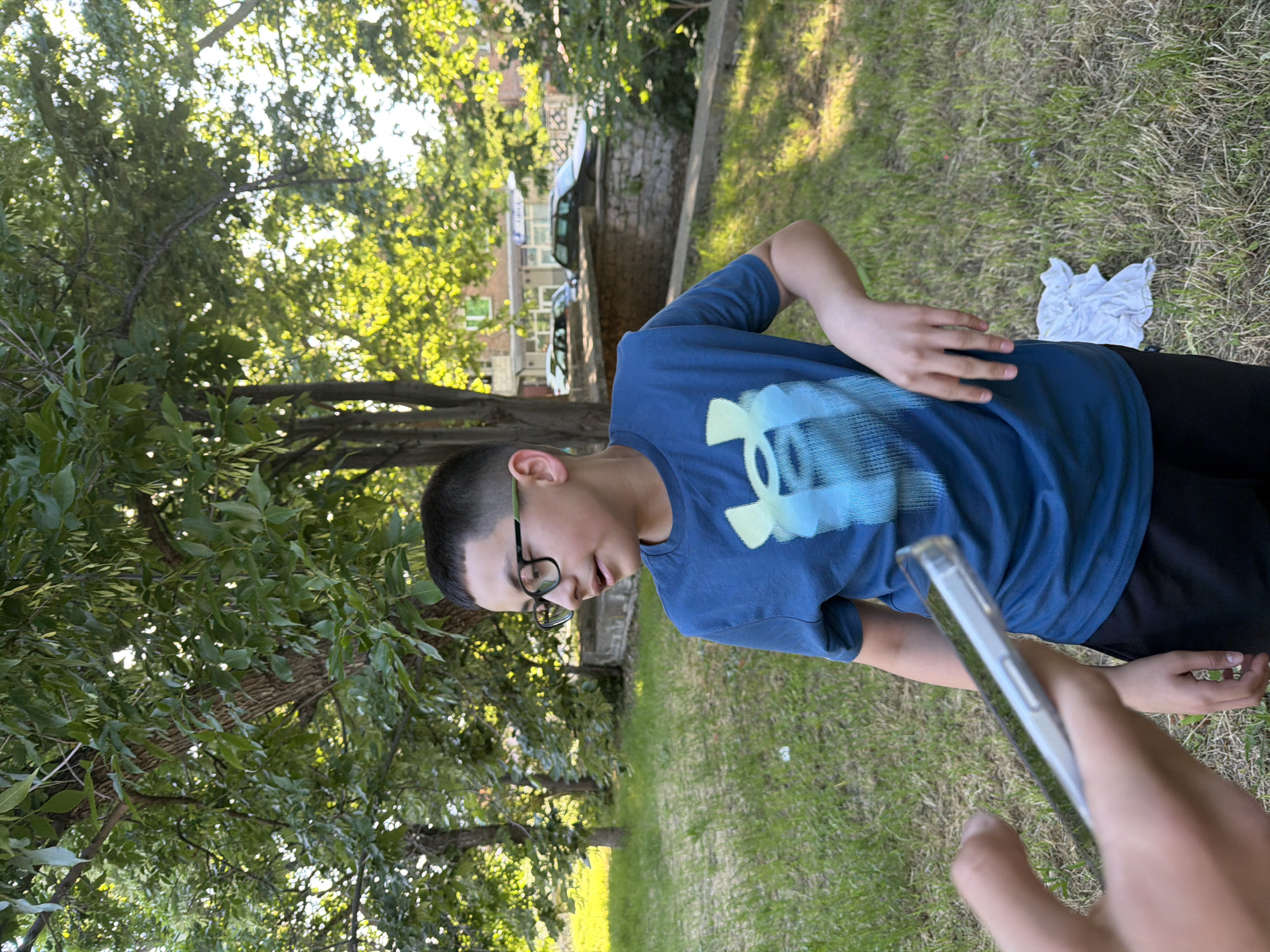 A teenager wearing glasses and an Under Armour t-shirt is standing on a grassy area. There's a white cloth on the grass nearby. The photograph appears to be taken in a park, as trees and buildings can be seen in the background. The location, Asenovgrad, Bulgaria, is not discernible from the image. A hand holding a smartphone is visible in the foreground.
