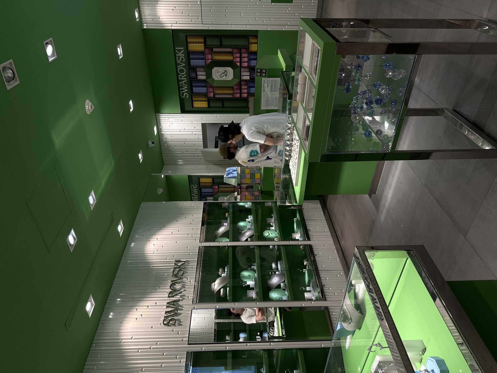 This image shows the interior of a Swarovski store, likely in Huixquilucan de Degollado, Mexico. The store features a modern design with green walls, a white, textured wall with "Swarovski" signage, and displays containing jewelry and decorative items. Two people are visible behind a jewelry counter. Display cases with green interiors showcase various products. Overhead lighting is visible in the green ceiling. The floor is tiled in a neutral gray.