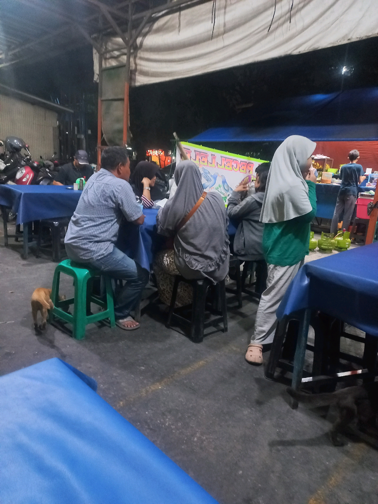 In Depok, Indonesia, this image captures an outdoor dining area at night, possibly a food stall or a small restaurant. Several people are seated at tables covered with blue tablecloths, engaged in dining or conversation. One man in a striped shirt and jeans sits on a green stool, while a woman in a grey hijab is seated at the same table. Another woman, also in a hijab, stands near the table. A young boy in a grey hoodie is also present. In the background, motorcycles are parked, suggesting a public area. The environment is dimly lit, with the presence of fluorescent lights and potentially string lights, creating a casual evening ambiance. A small, light-brown animal, possibly a dog, is visible near the man on the green stool. The ground appears to be paved with concrete. There are also large green gas cylinders near a food counter in the background. The overall scene depicts a typical evening gathering at an informal eatery.