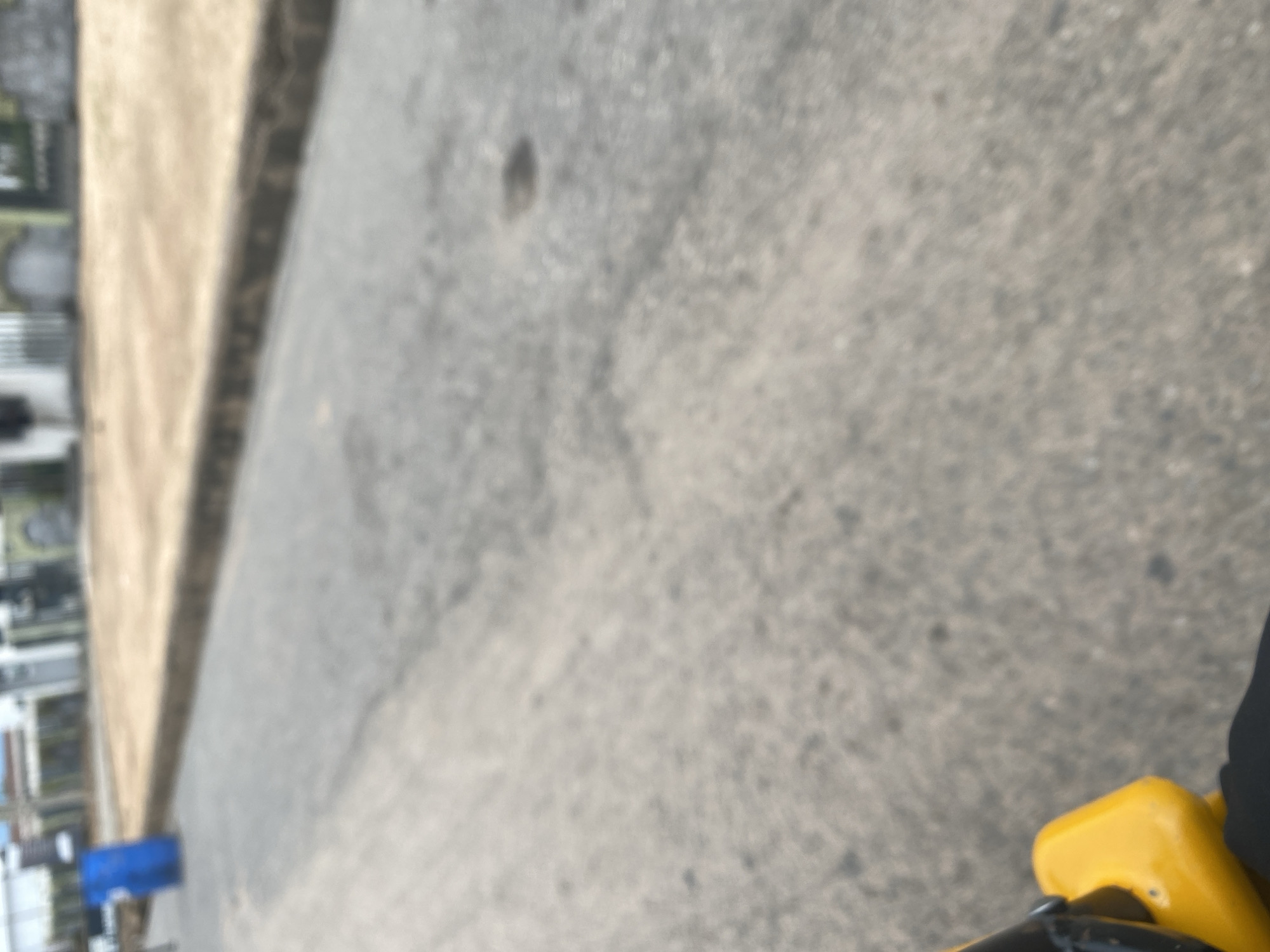 The image depicts a close-up, blurred view of a street scene in Jalingo, Nigeria. The perspective suggests the photo was taken from a moving vehicle or while holding a camera low to the ground, possibly from a bicycle.
The primary subjects are the street and its immediate surroundings. The ground is paved with asphalt, exhibiting a rough texture and scattered dark spots. To the left, a lighter-colored, slightly elevated strip of land runs parallel to the road, possibly a sidewalk or raised embankment. Further left, faint outlines of structures or objects are visible, appearing as vertical lines and rectangular shapes, suggesting buildings or parked vehicles, along with a bright blue rectangular object that might be a bin or container.
A small section of a bright yellow object, possibly a part of a vehicle or equipment, is visible in the lower right corner of the frame. The blurriness of the image makes it difficult to identify specific details about the time of day or weather, but the lighting does not suggest extreme darkness or bright sunlight, pointing towards a possibility of overcast conditions or a time other than midday. There is no visible text in the image. The overall impression is of an everyday street view, captured with a dynamic perspective that emphasizes motion and the immediate environment.
