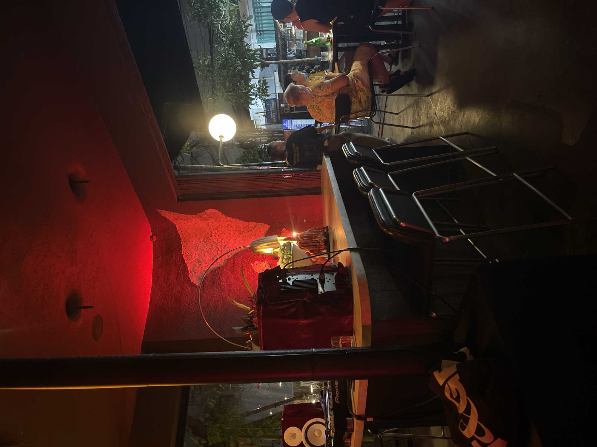 The image depicts the interior of a dimly lit bar in Mengwi, Indonesia, with a vibrant red light illuminating the textured wall and ceiling. In the foreground, a DJ setup with Pioneer speakers and a mixer is visible on the bar. A red velvet-covered structure, possibly a DJ booth or decorative element, sits on the counter, with a glass and a bottle of liquid nearby. Bar stools with metal frames and dark seats are arranged along the counter.
Through an open doorway to the right, the exterior street scene of Mengwi is partially visible, with trees, buildings, and streetlights. Several patrons are seated at tables outside, engaged in conversation and enjoying drinks. One person is standing near the bar, appearing to interact with someone behind the counter. A spherical light fixture hangs from a pole, casting a bright glow. The overall ambiance suggests a lively evening at a bar.