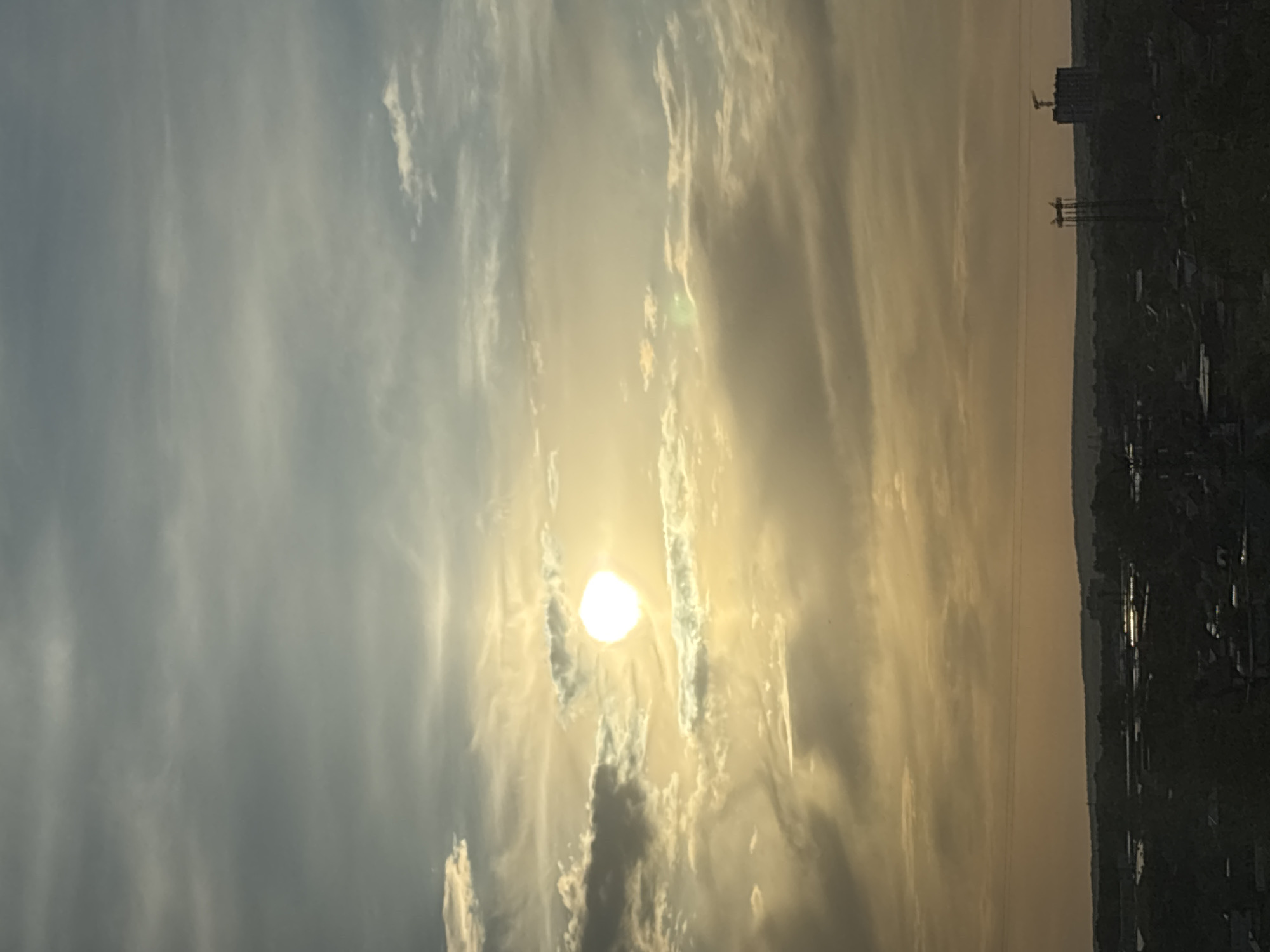 The image depicts a sunrise or sunset over the city of Chaikovsky, Russia. The sun is positioned in the center, partially obscured by clouds. The sky exhibits a gradient of colors, from pale blues and grays to warm yellows and oranges. A cityscape silhouette is visible on the right side, including buildings, construction cranes, and power lines.