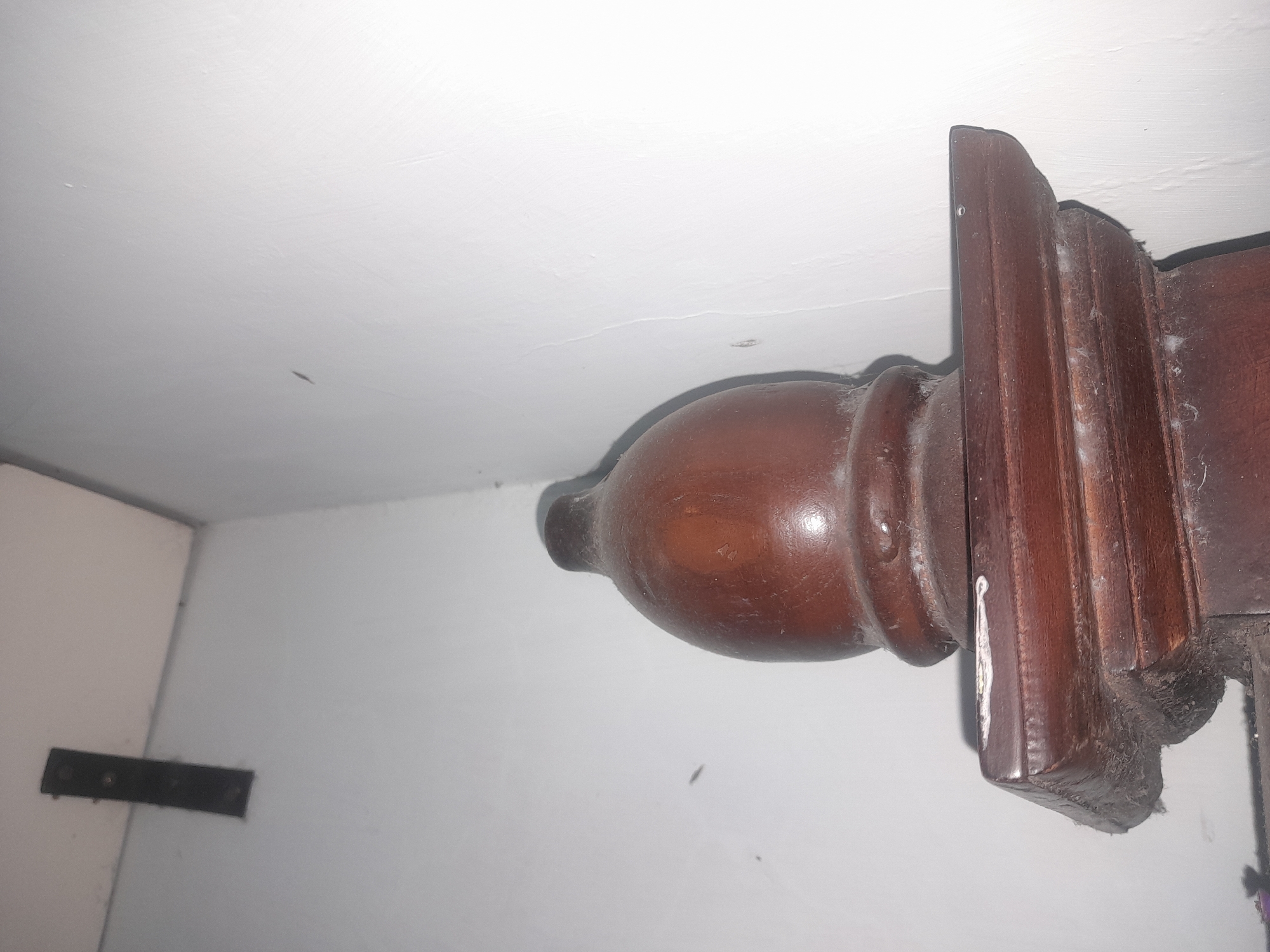 A close-up, low-angle interior shot captures the top of a dark wooden decorative post. The post features a rounded, bulbous top and a fluted lower section. It sits on a flat, rectangular wooden surface with layered, decorative edges. The post and surface are covered in a thin layer of dust and what appears to be cobwebs, indicating a lack of recent cleaning. The background consists of a light-colored wall that meets at a corner, with a dark rectangular object, possibly a bracket, mounted on the wall in the upper left quadrant. The lighting is dim, casting shadows and highlighting the textures of the dust and wood grain. The scene appears to be located indoors, within a residential or possibly older commercial space in Keboharan, Indonesia. There are no people or animals present.