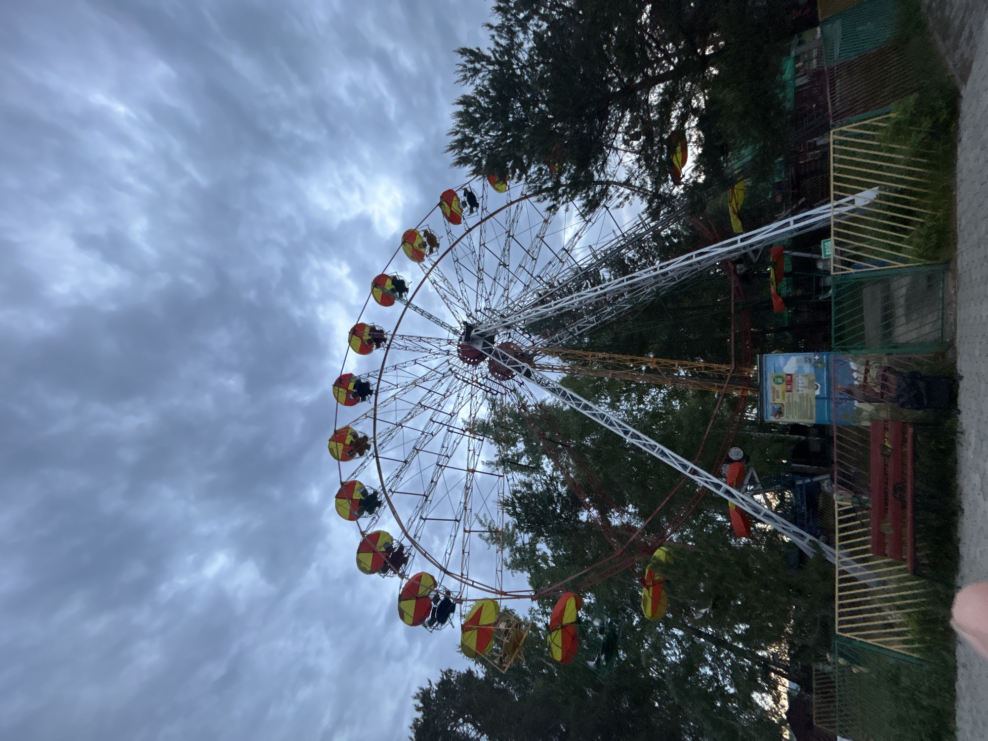 A Ferris wheel with red and yellow gondolas stands prominently against a backdrop of trees and a cloudy sky. The wheel appears to be located in a park or amusement area in the city of Chaikovsky, Russia. Adjacent to the wheel is a fenced enclosure with various signs and structures, including a sign with a map or diagram. The atmosphere is overcast, suggesting the possibility of rain. The wheel is partially obscured by trees in the mid-ground and the entire image is in landscape orientation.