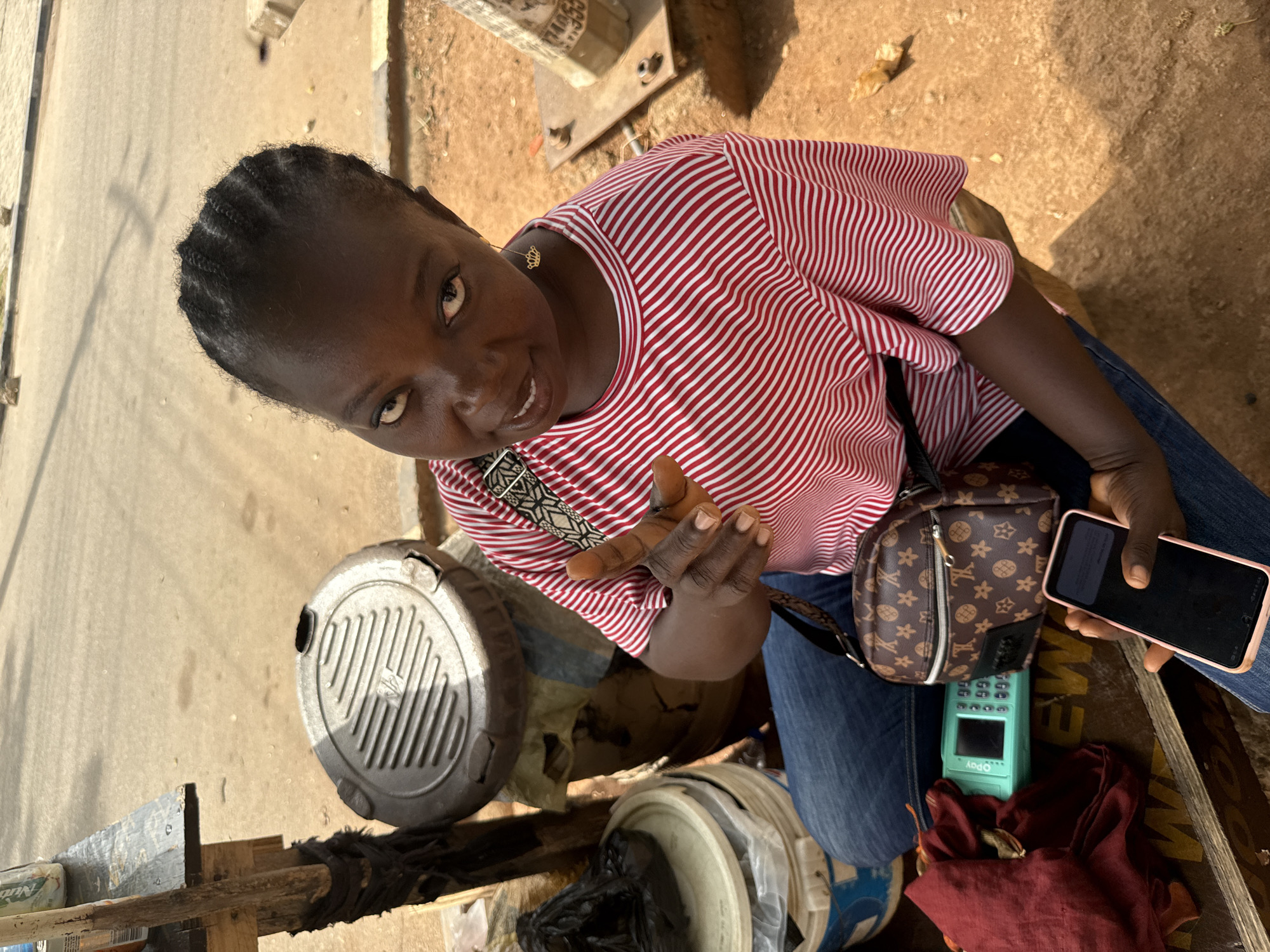 A high-angle outdoor shot captures a dark-skinned woman looking directly at the camera with a slight smile. She has braided hair and wears a red and white striped t-shirt, blue jeans, and a gold crown-shaped earring on her left ear. Her left hand is raised with fingers splayed, and she holds a black smartphone with a pink case in her right hand, its screen dark.
She appears to be seated or crouching behind a simple wooden setup, possibly a market stall or roadside stand, which is partially visible at the bottom of the frame. The ground is earthy and light brown, indicative of an outdoor setting in a sunny climate. The bright, even lighting suggests it is daytime.
Several objects are arranged around her, hinting at a commercial activity. Resting on her lap or a surface next to her is a small brown purse with a monogram pattern. Next to it, a mint green Opay point-of-sale (POS) machine is visible. Below the machine is a piece of maroon fabric. To her left, two circular, possibly metallic or plastic, containers with lids are present, one large and gray with a gridded top, and another smaller, white one. A partially visible bar of soap with "Nutri" printed on it can be seen on the wooden structure to the bottom left. The overall scene suggests a vendor operating a mobile money or small retail business in an outdoor environment, common in places like Abuja, Nigeria.