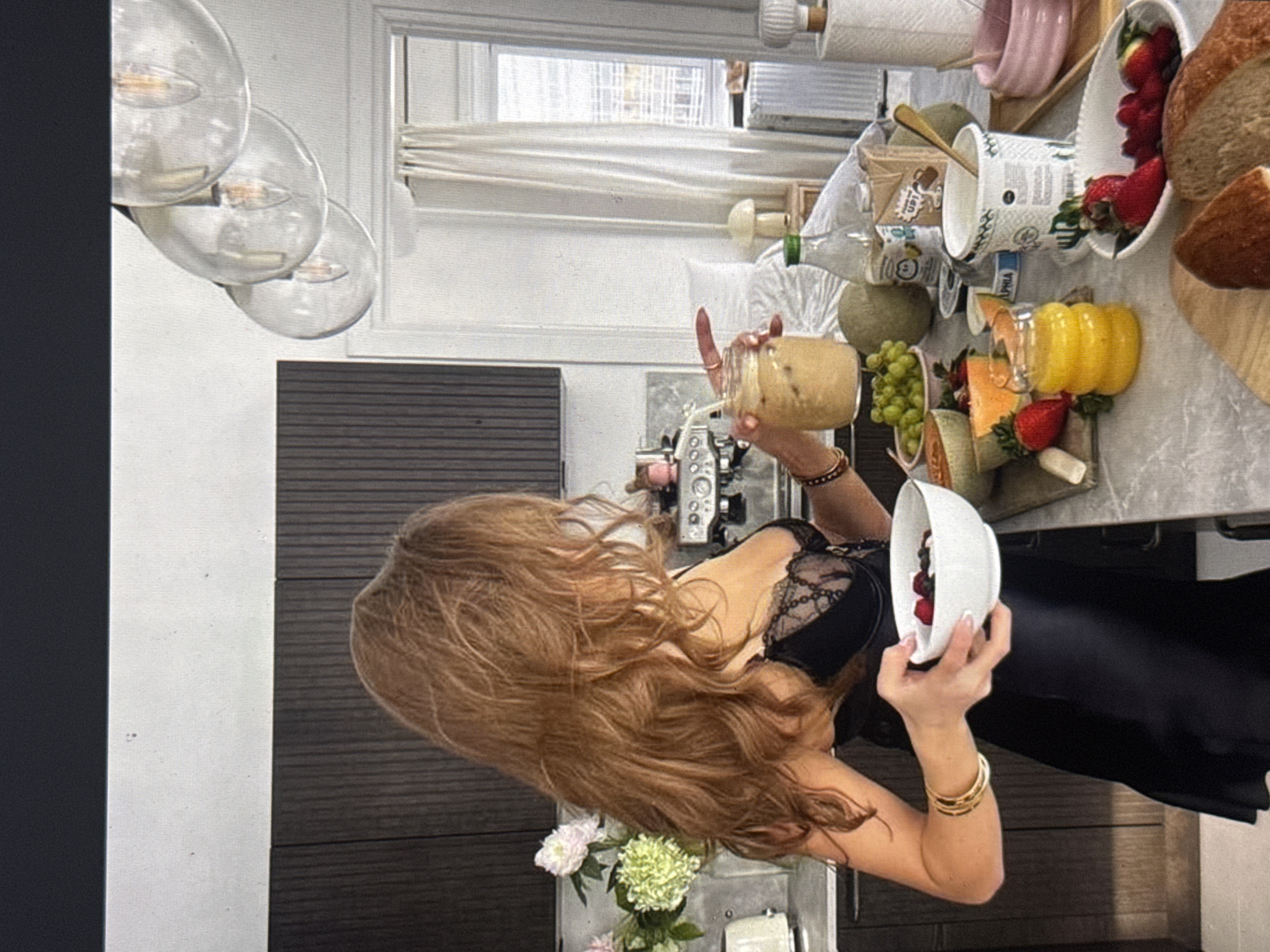 The image shows a woman with long, wavy auburn hair wearing a black lace-trimmed camisole and a black skirt. She is standing in a brightly lit kitchen with dark wood cabinetry. The woman is holding a glass jar filled with a light-colored beverage, possibly a smoothie or coffee drink, in her right hand. She is also holding a white bowl containing berries in her left hand.
On the counter, there is a variety of food items, including grapes, cantaloupe, bell peppers, strawberries, raspberries, and bread. There are also some reusable cups and a glass bottle. The overall scene suggests a healthy breakfast or snack preparation. The lighting and abundance of natural light imply it is daytime. The kitchen appears modern and well-equipped, with a window in the background. The woman's expression is not clearly visible, but her posture suggests she is engaged in preparing or enjoying her food. The image is a close-up, focusing on the woman and the food on the counter.