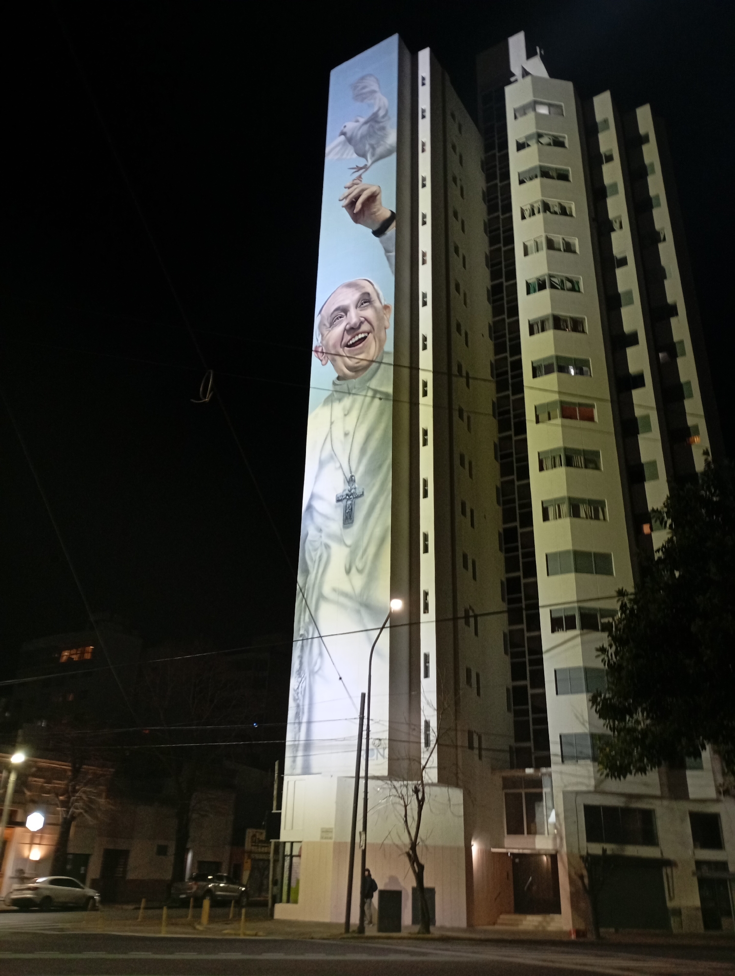 A nighttime street-level view captures a tall, multi-story building in La Plata, Argentina, adorned with a large mural of Pope Francis. The mural, illuminated by an unseen light source, covers a significant portion of the building's facade. It depicts Pope Francis from the chest up, wearing a white cassock and a large silver cross necklace. His head is tilted back, and he is smiling broadly. Above his head, a white dove is depicted in flight, with a hand extended from off-frame holding it. The building itself is contemporary in design, with multiple windows and balconies. The street in front of the building is visible, with cars parked and driving by. Streetlights cast a warm glow on the scene, and trees line the sidewalk. The overall impression is one of urban life at night, with art integrated into the cityscape.