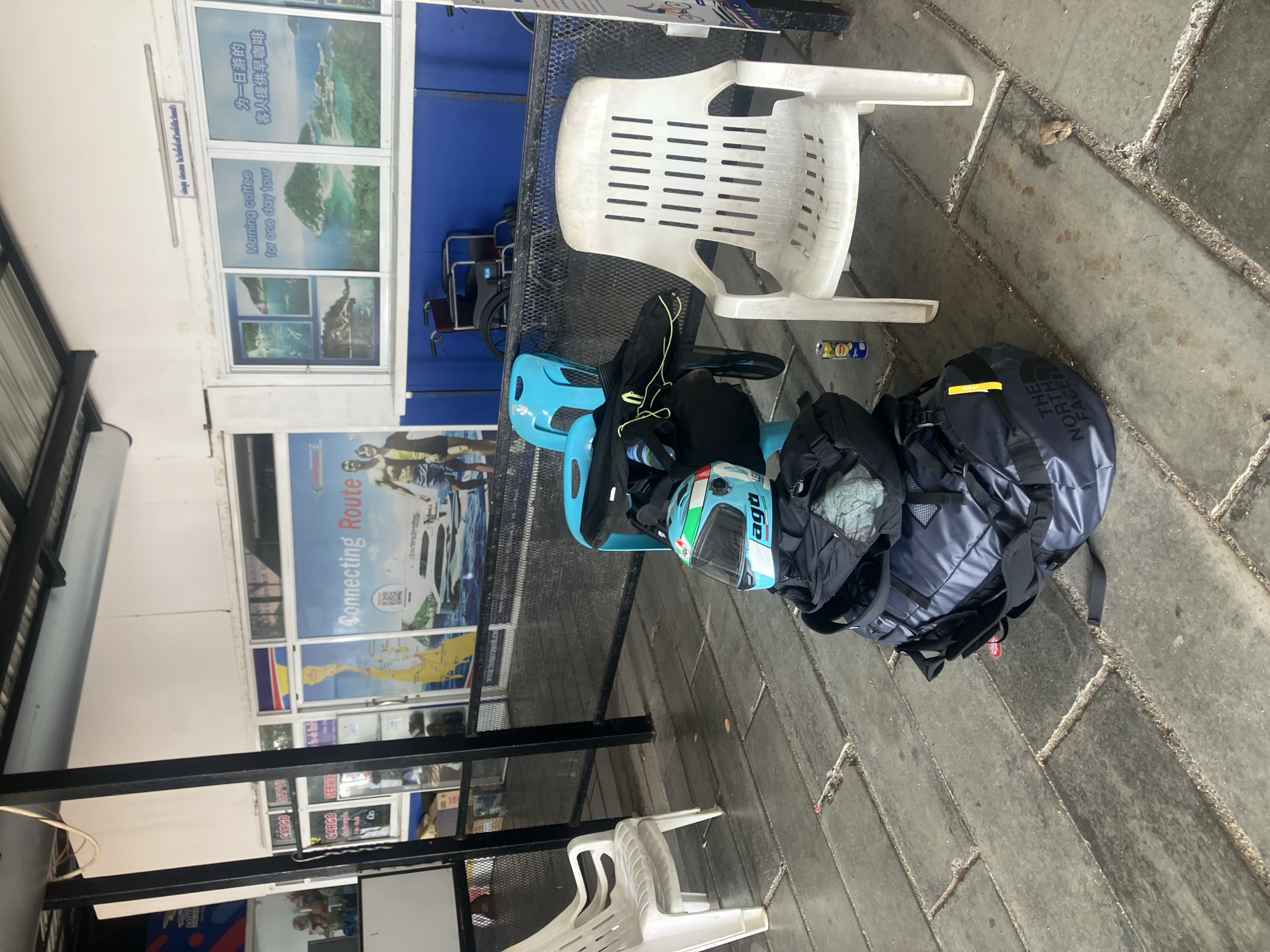 The image depicts an outdoor scene in Baan Mae Nam, Thailand. A blue motorcycle helmet and a white AGV helmet are placed on a light blue plastic chair. A black backpack and a navy blue The North Face backpack are placed next to the chair. A white plastic chair is positioned next to the helmets and backpacks. The scene is set against a backdrop of windows displaying tourism-related posters and text in both Thai and English. The ground is paved with grey stone tiles. The scene appears to be near a travel agency offering tours and cargo services.