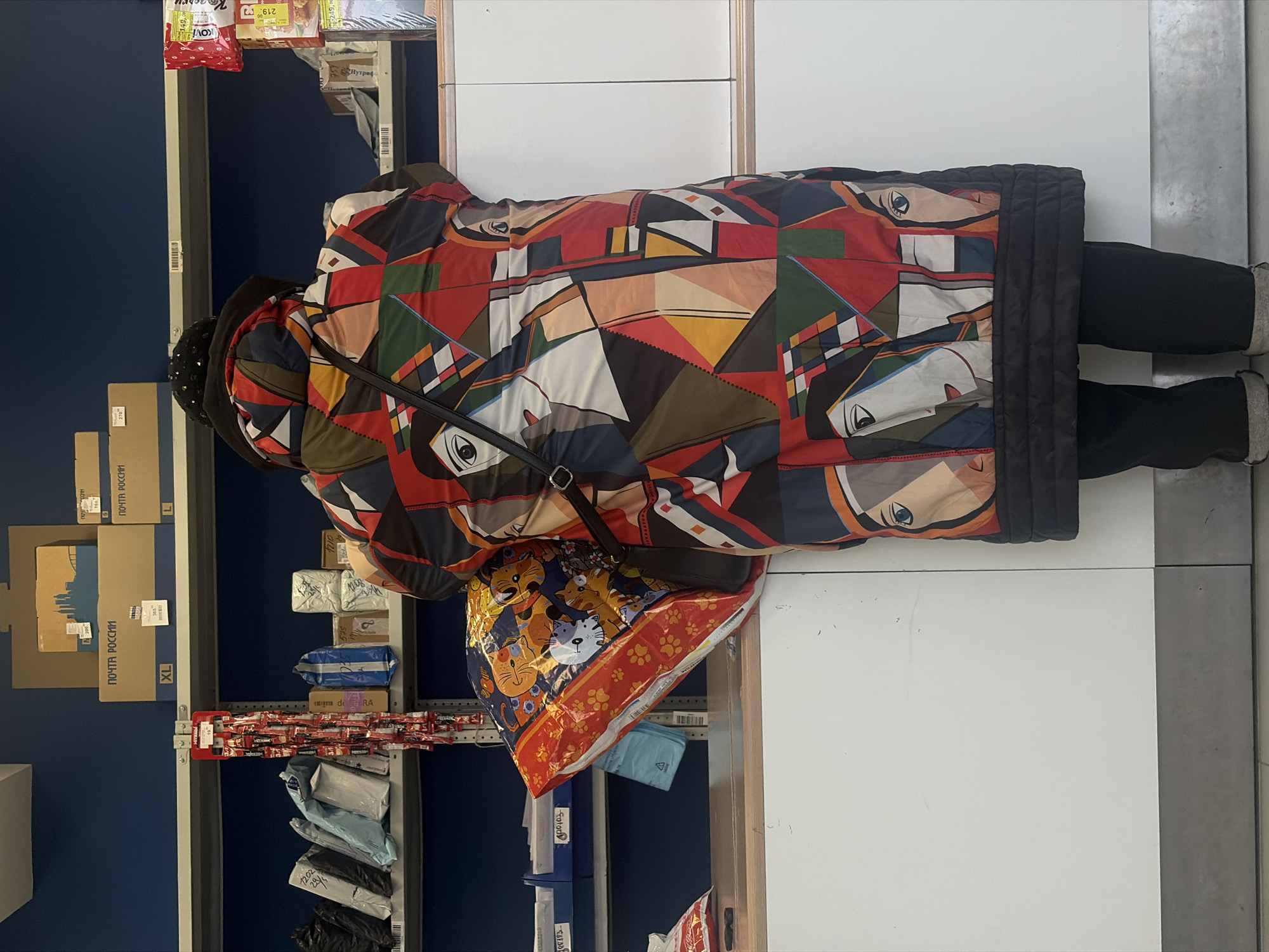 The image captures a person from behind within what appears to be a Russian Post office in Saint Petersburg, given the "ПОЧТА РОССИИ" (Russian Post) labeling on visible boxes and the general setting. The person is wearing a distinctive, colorful winter coat with an abstract, geometric pattern featuring stylized faces in shades of red, blue, green, yellow, and white. They are also dressed in black trousers and light-colored shoes or boots, carrying a black shoulder bag. In their arms, they hold a large, vibrant bag decorated with cartoon cats, which appears to be packaging for a pillow or plush toy.
The scene is indoors, characterized by dark blue walls and metal shelving units on both sides of the person. These shelves are stocked with various items, including cardboard shipping boxes (some marked "XL" and "L"), packaged mail parcels in blue, white, and black wrapping, and smaller retail items. One shelf prominently displays numerous small red packets, partially revealing "NESCAFÉ" branding, suggesting snacks or beverages for sale.
Visible text from OCR includes "ПОЧТА РОССИИ," sizes "XL" and "L" on boxes, and various numbers such as "1202," "270," "365," "219," and "249," which likely denote parcel numbers, prices, or product codes. Partial brand names like "KOVI" and "Нутриф" (Nutrif) are also discernable. The heavy winter coat worn by the individual suggests the image was taken during colder months. The activity appears to be consistent with someone waiting or conducting a transaction at a postal service counter, although the counter itself is not fully visible.