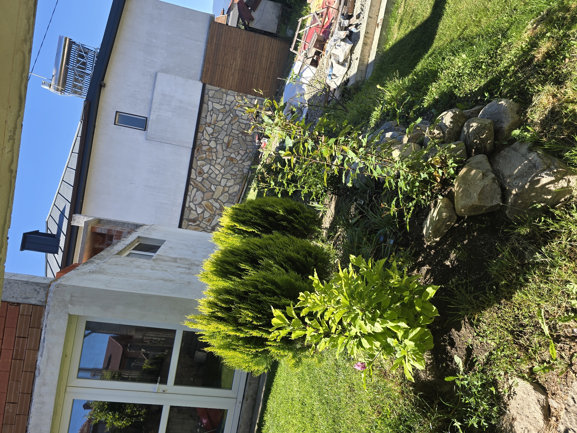 The image depicts a garden scene in Dorkovo, Bulgaria, adjacent to a two-story building. The building features a combination of white stucco and decorative stone veneer on its exterior, and it has a solar water heater on the roof. A pair of white-framed windows with reflective glass are visible on the ground floor.
The garden contains several plants, including two dense, dark green shrubs and a taller, leafy plant with slender branches. A rockery is located near the property line. The lawn is patchy with varying shades of green. A wooden fence is partially visible in the background.