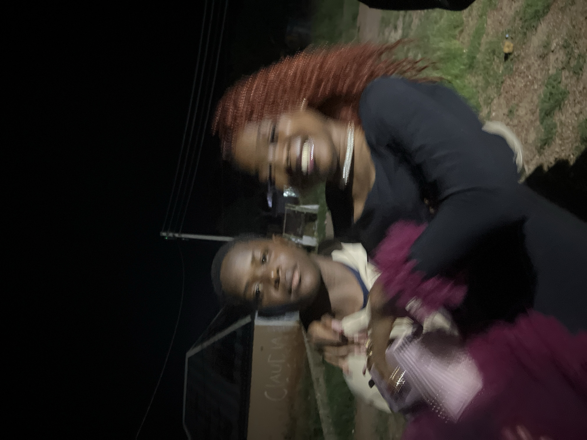 In Durumi, Nigeria, at night, a young Black male stands in the foreground, his face turned towards the camera and his gaze direct. He wears a light-colored jacket over a dark shirt. To his right, a young Black female with vibrant red hair is partially visible, smiling broadly and looking away from the camera. She has a shimmering necklace around her neck. Her arms are around the male, and she is holding a pink object, possibly a purse, with both hands. Their interaction suggests a close relationship, possibly romantic or familial. The background is dark, indicating it is nighttime, with a building visible to the left, possibly a house, on which the word "CLAUDIA" is scrawled in white. A utility pole with wires is present to the right. The ground is covered in dirt and patches of grass. The overall scene appears to be an outdoor gathering or casual encounter.