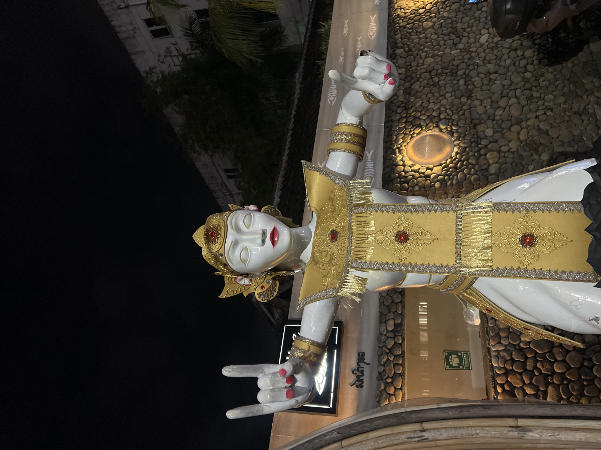 A striking statue of a stylized female figure, possibly a deity or traditional dancer, stands prominently in an outdoor setting at night. The statue is predominantly white, adorned with intricate gold and red embellishments, including a golden headdress with floral accents and a gold and red ornate top with matching armbands. Her face is white with closed eyes and red lips, conveying a serene expression.
Notably, her hands display contrasting gestures: the left hand forms a "rock on" sign (index and pinky fingers extended), while the right hand has curled fingers with the index finger extended. Both hands feature red nail polish. The ground around the statue is covered with small, round, natural-colored pebbles, and a warm-toned ground light illuminates a section of the pebbles.
In the background, a dark night sky contrasts with a dimly lit building, partially obscured by sparse tropical foliage, including palm fronds. A motorcycle or scooter is partially visible to the far right. Several details adorn the surroundings: a vertical pattern of stylized fish bones is visible on a light-colored wall section. Below the statue's left hand, a dark sign with a white border displays stylized text, possibly "Rock Star," and local script (likely Balinese or Indonesian, appearing as "Satyagraha" or similar) is seen further to the right. A green emergency exit sign with pictograms is also visible against a light-colored wall section on the right. The overall scene suggests an establishment in North Kuta, Indonesia, incorporating traditional aesthetics with a modern, perhaps playful, touch.