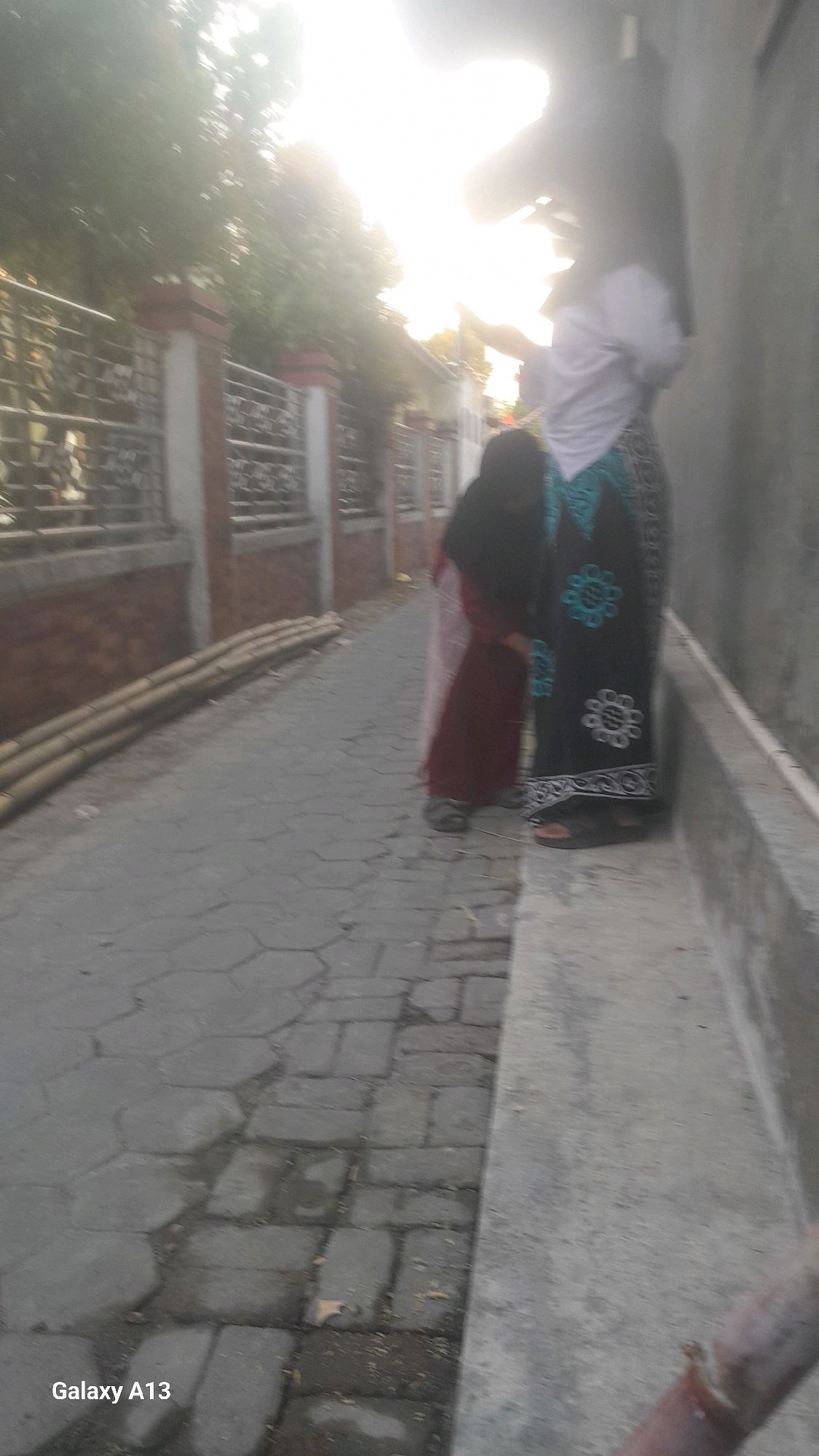The image, taken in the city of Ngelo, Indonesia, captures a sunlit alleyway. In the foreground, a person wearing a dark hijab and a maroon dress stands next to another individual dressed in a white shirt and a black skirt with a blue floral pattern. To the left, a low fence with decorative metalwork separates the alley from a building with red-brick accents. Stacked bamboo poles are visible along the fence. The ground of the alley is paved with hexagonal grey stones, and the overall lighting suggests it is either early morning or late afternoon. The lower left corner of the image displays "Galaxy A13" indicating the device used for capture.