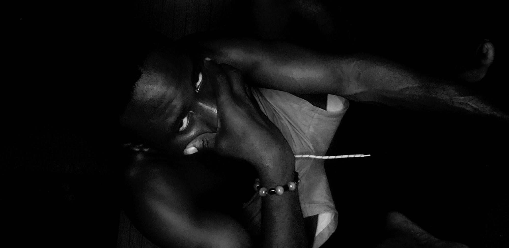 The image is a black and white photograph of a dark-skinned person with light-colored eyes. The person is lying down with their head resting on their hand, a bracelet is visible on their wrist. Part of the person’s torso is visible, showing the edge of a white garment. A thin white object is adjacent to the garment. The background is dark and indistinct. There is no information in the image to verify the location as Narayi, Nigeria.