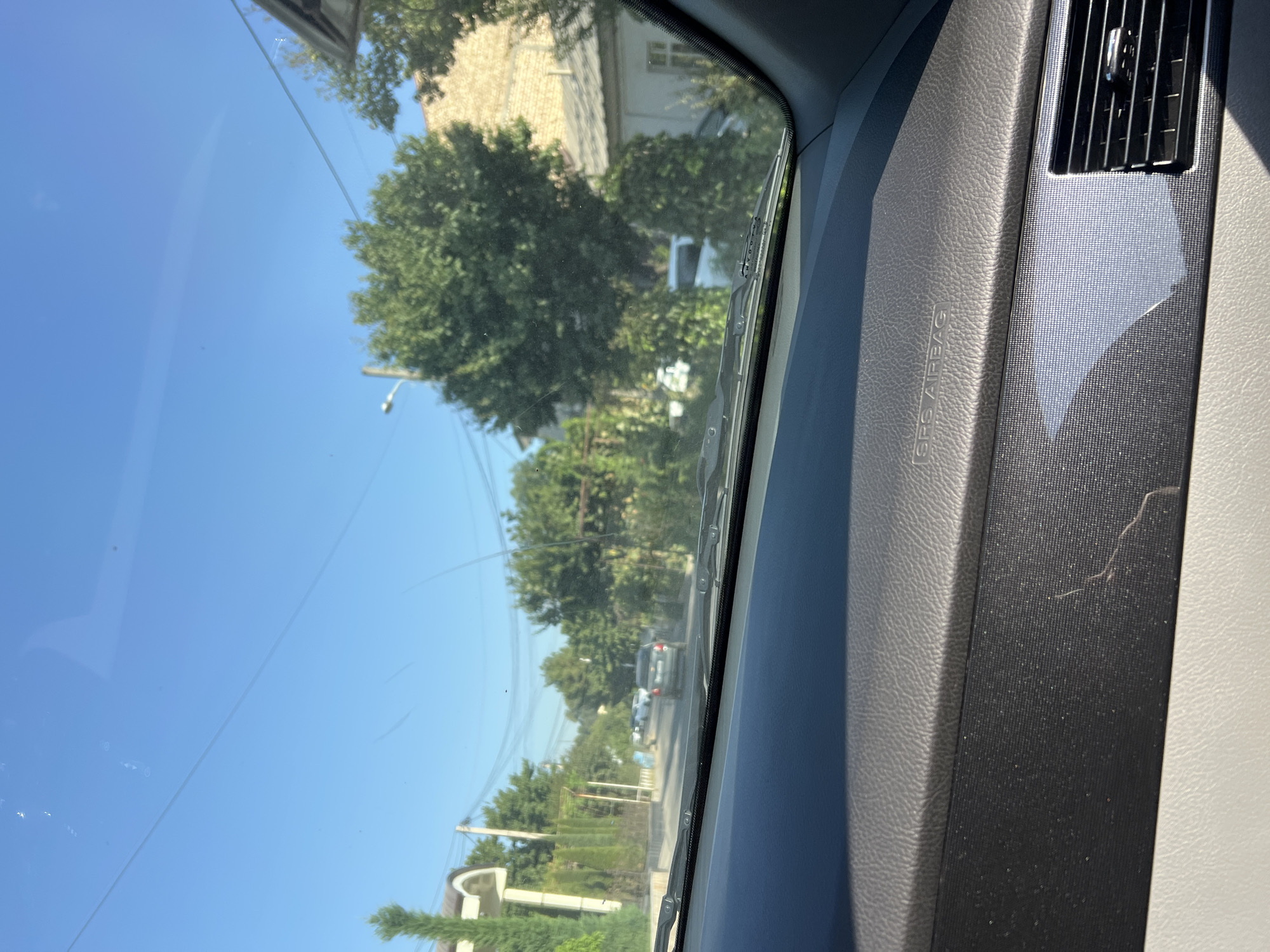 The image is taken from the interior of a car, looking out through the windshield. The view shows a street scene with trees, houses, and other vehicles. The sky is clear and blue. The car's dashboard is visible in the foreground, with the "SRS AIRBAG" label clearly printed on it. The street appears to be in a residential area. The time of day seems to be daytime, judging by the bright sunlight. There are no people visible in the image.