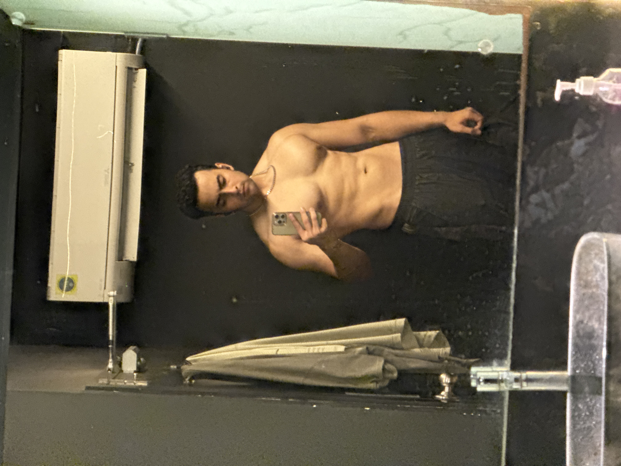 A man is taking a mirror selfie in what appears to be a gym or bathroom. He is shirtless, displaying his physique, and wearing dark athletic shorts. In the background, an air conditioner unit is mounted on the wall, and a towel is hanging on a hook. The mirror reflects the man and the surrounding environment, including a sink and faucet in the foreground. The image appears to be taken in an indoor setting. There is no information in the image to suggest a location in Kolkata, India.