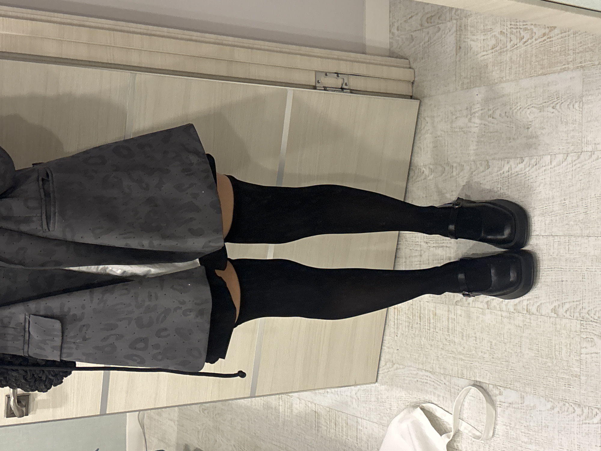 The image, captured in an indoor setting and presented rotated 90 degrees clockwise, focuses on the lower body of a person. The subject is wearing a dark gray garment, possibly a skirt or jacket, which features a subtle, tone-on-tone leopard or abstract spot pattern and a visible flap pocket on the left side. Beneath this garment, the person wears opaque black tights. Their feet are adorned with black, chunky Mary Jane style shoes, characterized by thick soles and buckle fastenings across the instep.
The setting consists of a light-colored wooden floor made of wide planks, providing a clean and bright base. In the background, there is a large, light-colored wooden surface, which appears to be part of a wardrobe or a folding door, indicated by a visible hinge mechanism near the top edge.
Additional objects in the scene include a black bag or purse, partially visible on the far left next to the subject's leg, and a white fabric tote bag with handles, lying on the wooden floor near the subject's feet on the right. The lighting is consistent with an indoor environment, offering no specific cues regarding the time of day or outdoor conditions. There is no visible text on any items in the image. The scene is presented as a casual, possibly posed, shot of an outfit, located in Krasnoyarsk, Russia.