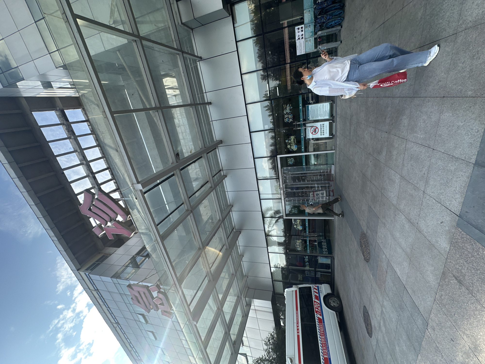 The image shows the exterior of the Outpatient Department of the First Affiliated Hospital of Chongqing Medical University, located in the High-tech Zone of Chongqing, China. The building features a modern facade with large glass windows and an overhead glass canopy. Prominent red Chinese characters reading "急诊" (Emergency) are visible on the upper portion of the building.
In the foreground, a white ambulance with red and blue stripes is parked to the left. The ambulance has Chinese text on its side, including "重庆医科大学附属第一医院" (First Affiliated Hospital of Chongqing Medical University) and "120" which is the emergency number in China.
A woman is walking in front of the building's entrance, looking at her phone. She is wearing a white t-shirt, a light blue striped shirt, and blue jeans. She is holding a red shopping bag. To her left, a man is walking towards the entrance.
The ground is paved with grey tiles. The sky above is blue with some clouds, and the sun is casting shadows and reflections on the building. Bicycles are parked near the entrance on the right side. Several signs with Chinese text are posted near the entrance doors, one of which appears to be a no-smoking sign.