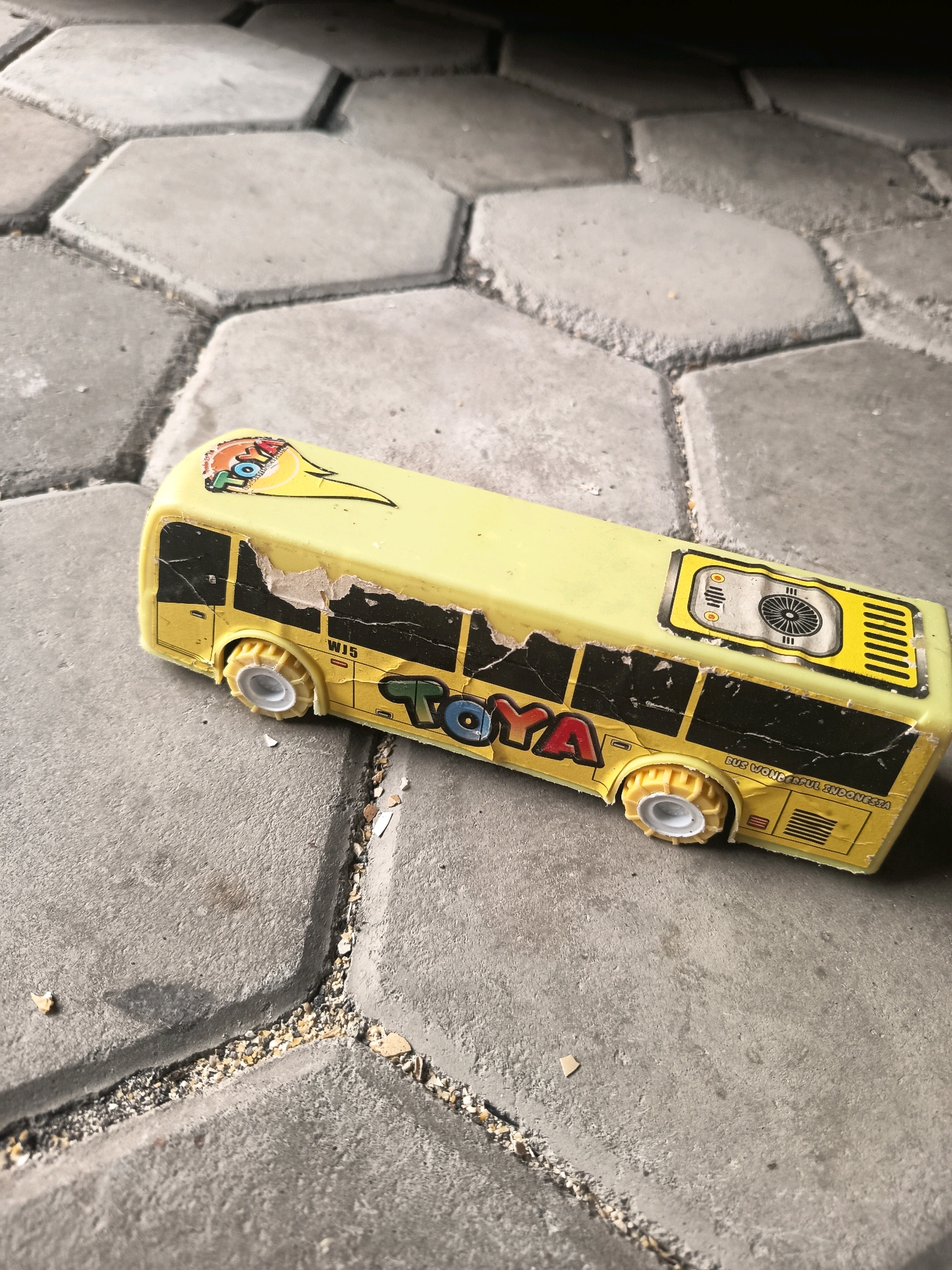 A yellow toy bus is lying on a gray concrete hexagonal pavement in Sidorejo, Indonesia. The bus is worn, with peeling stickers and damaged paint. The stickers on the bus display the word "TOYA" and other graphics. The pavement is cracked and shows signs of wear and tear, with small pebbles and debris scattered in the cracks. The lighting suggests it is daytime. There are no people visible in the image.
