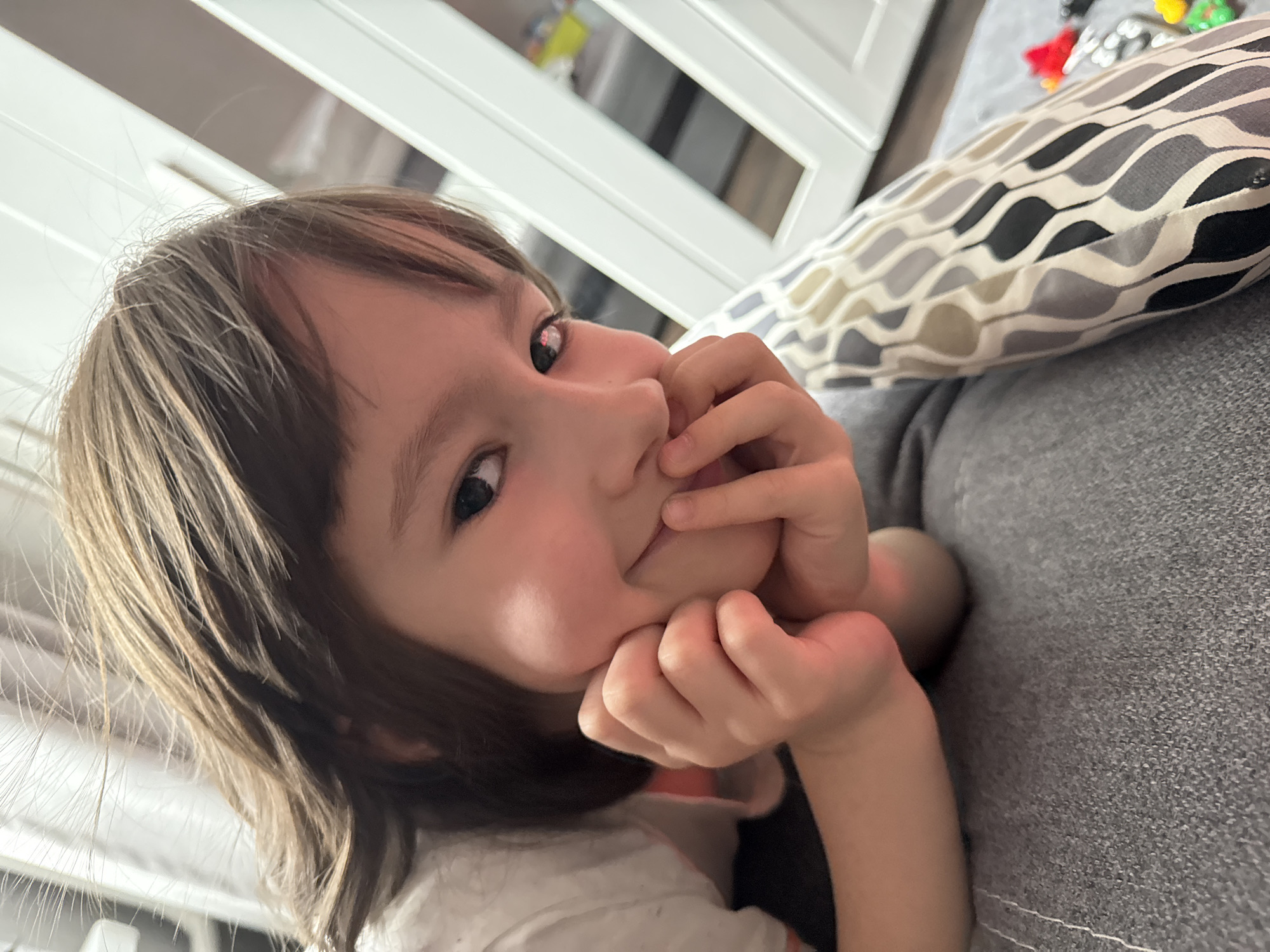 A young child with light brown hair and dark eyes is depicted in a close-up, tilted portrait. The child's face is partially framed by their hands; one hand has a finger resting on the lips, while the other supports the chin. A gentle, curious, or slightly shy expression is visible as they look directly towards the viewer. Reflections of a white, possibly rectangular light source are visible in their eyes, suggesting an indoor setting during the daytime.
The child is reclining on a grey, textured upholstered surface, likely a couch or bed. Adjacent to them is a decorative pillow featuring a wavy, geometric pattern in varying shades of grey, black, and light beige. In the blurred background, a white furniture piece with horizontal slats or panels can be seen, along with indistinct, colorful objects (red, yellow, green) that appear to be toys. The overall scene conveys an intimate, candid moment indoors. No text is visible in the image.