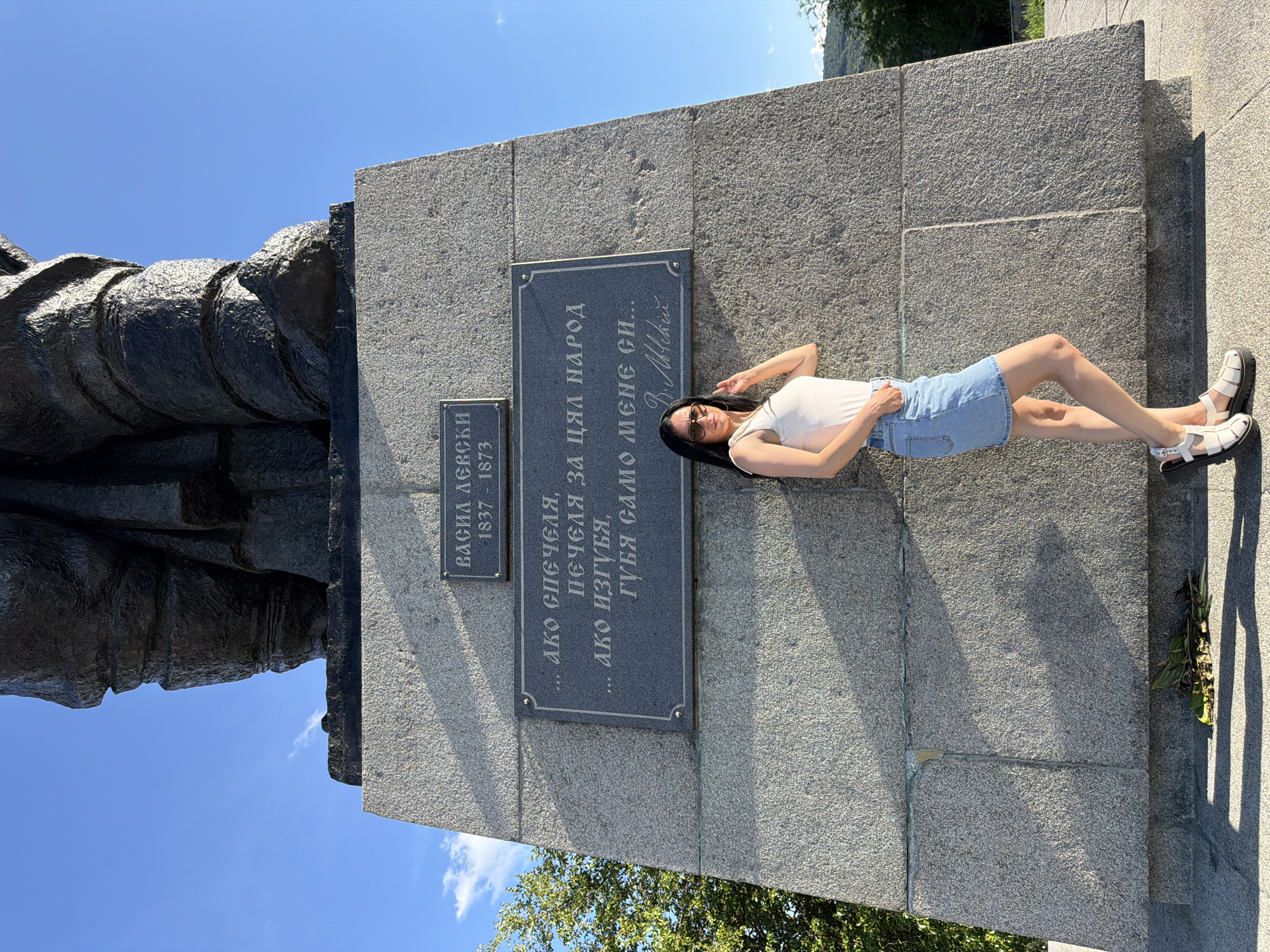 A medium shot shows a woman posing in front of a monument. The monument is made of rough, grey stone blocks and features a large bronze statue on top. Two plaques are visible on the monument's face: one at the top reads "ВАСИЛ ЛЕВСКИ 1837 - 1873" (Vasil Levski 1837 - 1873), and below it, a larger plaque contains text in Bulgarian: "... Ако спечеля, печеля за цял народ ... ако изгубя, губя само мене си..." (... If I win, I win for the whole nation ... if I lose, I lose only myself...). The woman is wearing a white tank top, a light blue denim skirt, and white platform sandals. She has dark hair and is wearing sunglasses. She is leaning against the monument with one hand in her pocket and the other resting on her hip. The monument appears to be located in an outdoor setting with a blue sky, some trees on the left, and a hint of a hillside in the background on the right. The monument is the Vasil Levski Monument located in the town of Lovech, Bulgaria.