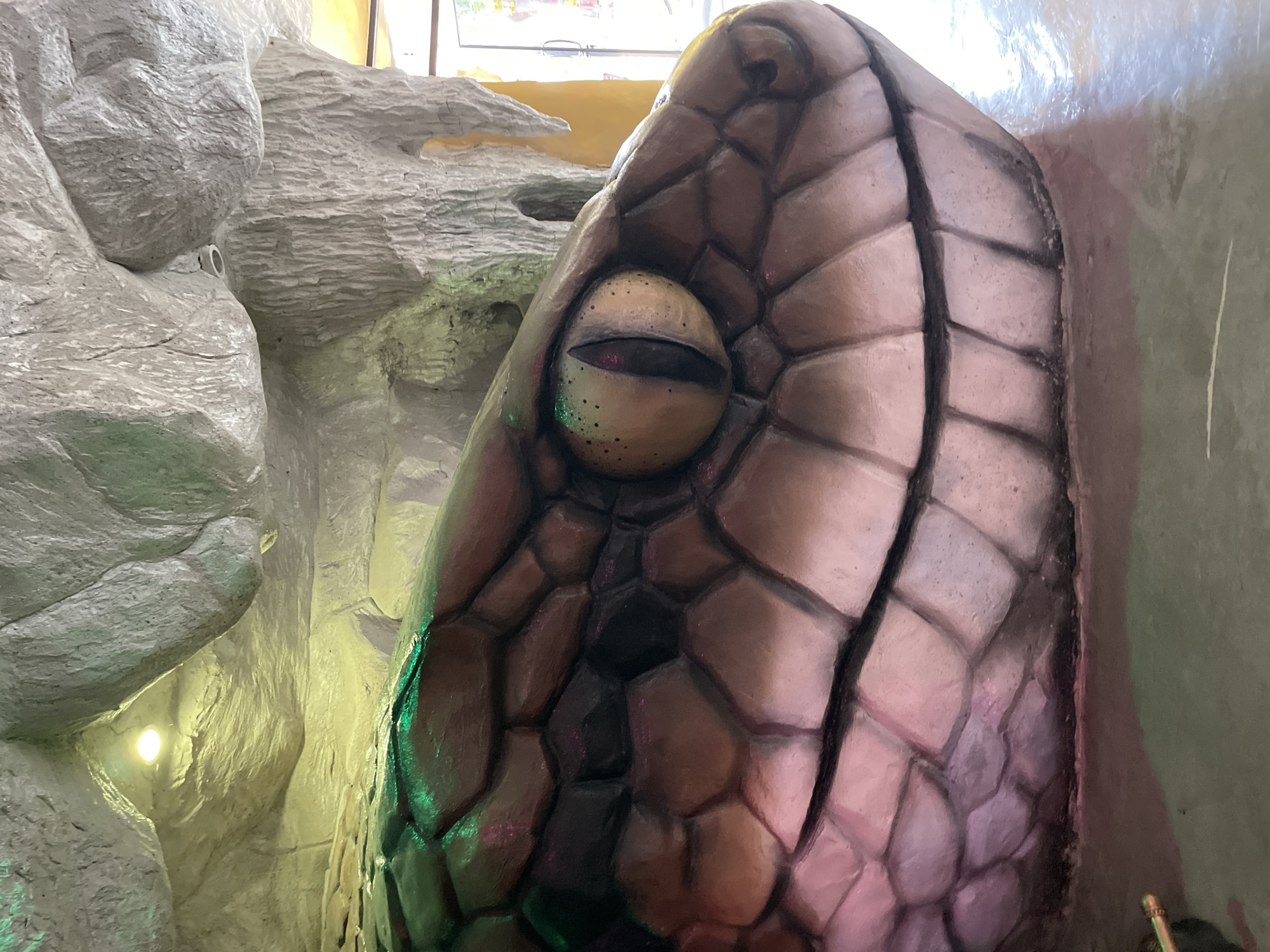 A close-up, eye-level shot of a large, artificial snake head sculpture is centered in the frame. The sculpture is highly detailed, with individual scales rendered in shades of brown, pink, and green, illuminated by ambient lighting. The snake's eye, with a slit pupil, is a prominent feature, reflecting a yellowish hue. The background consists of textured, cave-like rock formations, with visible lighting fixtures. The floor beneath the sculpture appears to be a glossy, light-colored surface. Part of a doorway and a yellow wall are visible to the right, suggesting the sculpture is located indoors, possibly within an attraction or themed environment in Bo Win Subdistrict, Thailand.