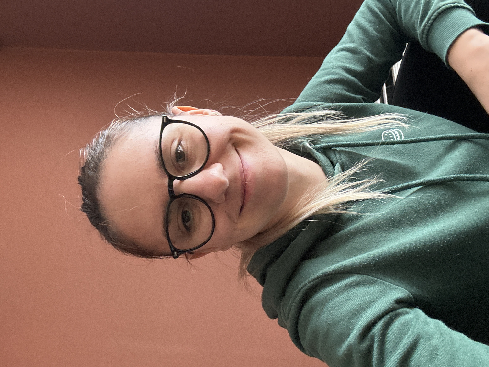 A woman with glasses and a green hoodie is shown in a selfie-style image. The background is a solid, light brown color, likely a wall. The location is not identifiable as Sofia, Bulgaria, based on the provided image. The focus is on the woman's face and upper body, and she is smiling slightly. Her hair is blonde and pulled back. A small portion of a black object is visible behind her shoulder.