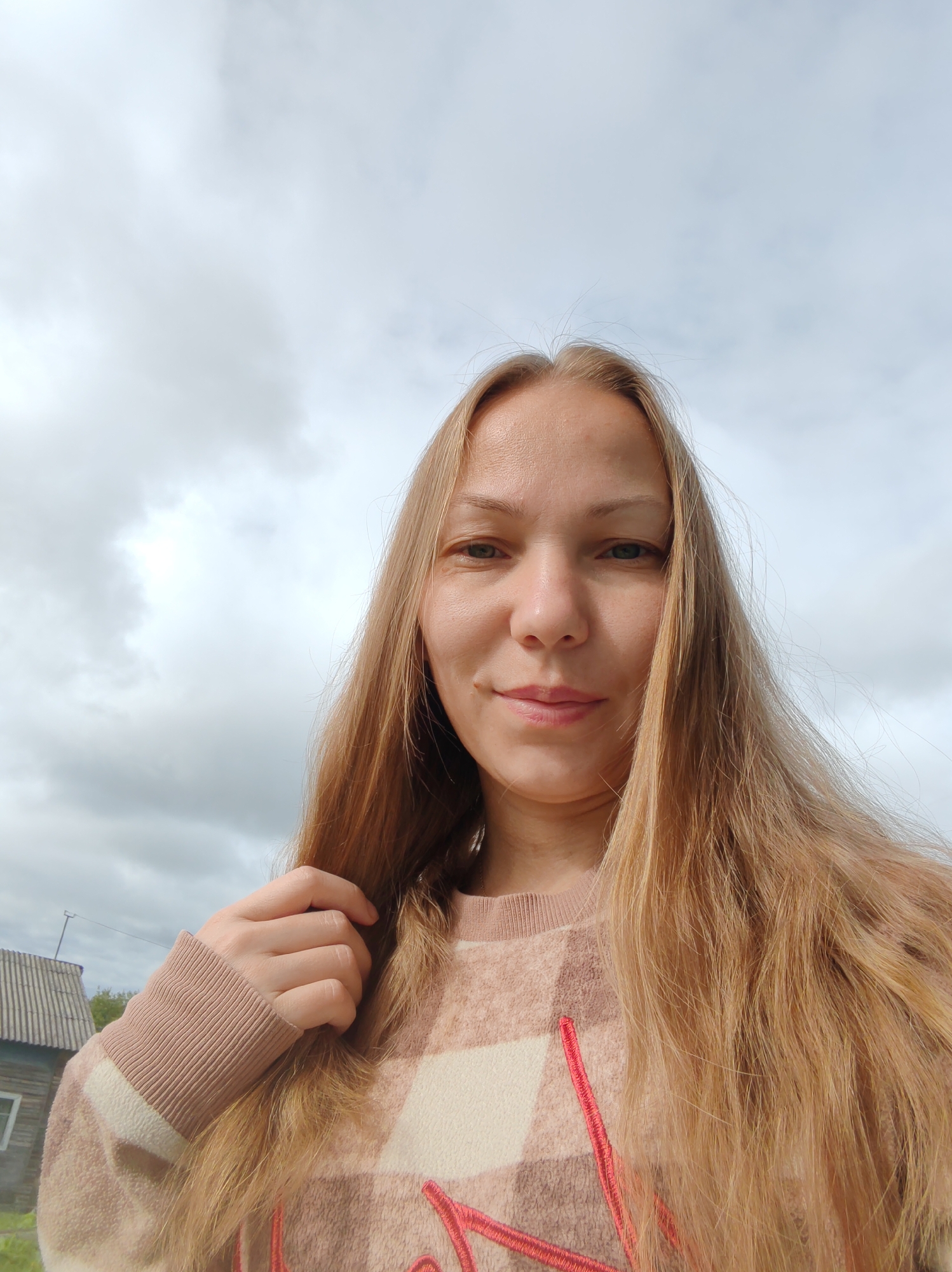 A young woman with long, blonde hair and light green eyes is pictured from the chest up, looking directly at the camera. She is wearing a beige and cream plaid sweater with a red embroidered detail. Her left hand is raised and touching her hair. In the background, a cloudy sky dominates the upper portion of the frame, while the lower left shows the corner of a simple, wooden house with a gray, corrugated roof, indicative of a rural or suburban setting. The overall environment suggests an outdoor scene, likely in the village of Chukhlem, Russia.