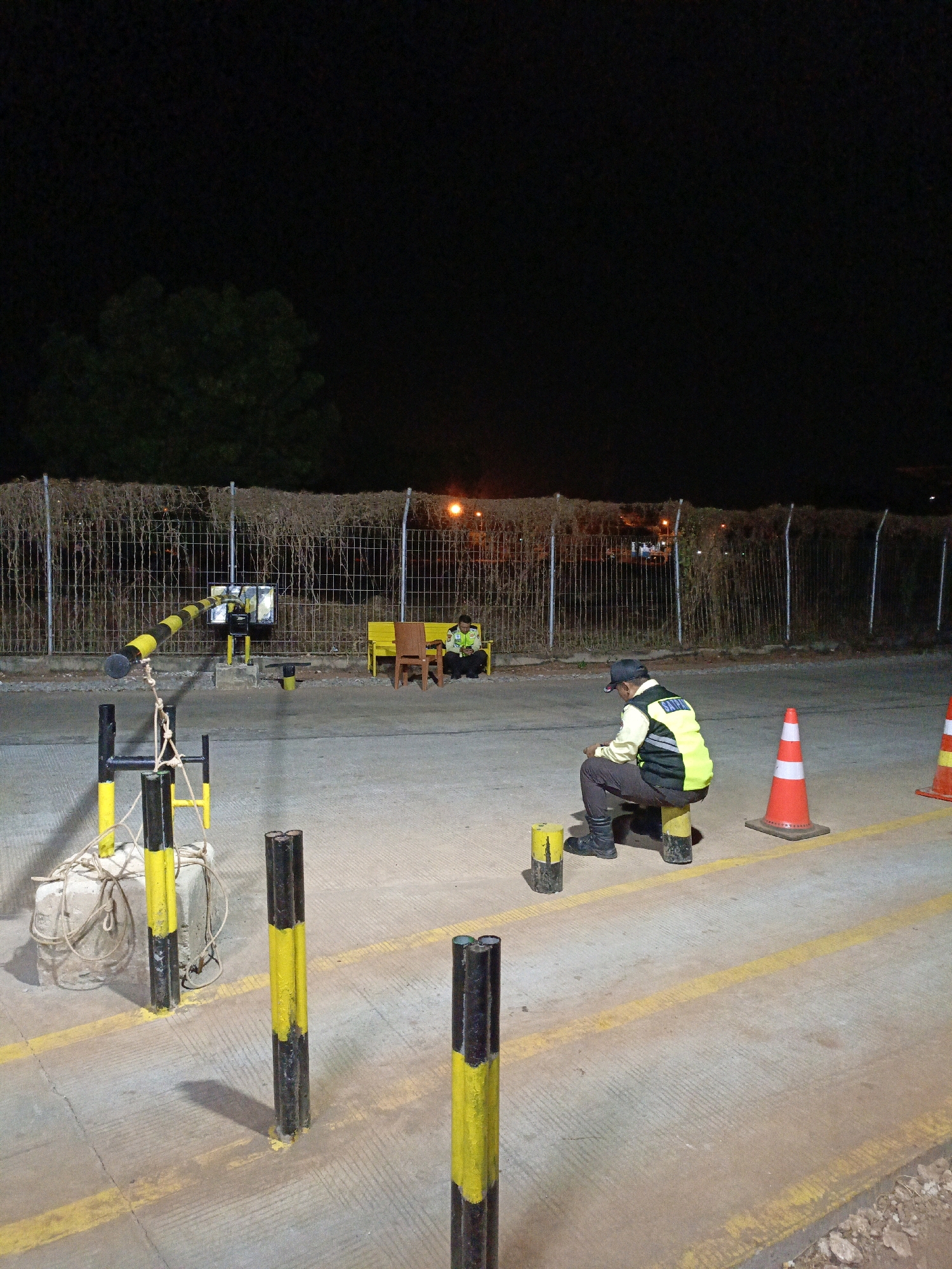 A night time photograph taken in Jenu, Indonesia shows two male security guards seated on short, yellow-topped posts on a concrete surface marked with yellow lines. The guard in the foreground, wearing a dark baseball cap, a light-colored shirt, and a bright yellow reflective vest over a darker vest, is looking down at his phone. He is seated on a post in front of two orange traffic cones and a yellow and black striped bollard. In the background, another security guard in similar attire is seated on a yellow bench next to a brown chair, also appearing to be looking at a device. Behind them, a metal fence covered in dry vegetation runs horizontally, with distant lights visible beyond it. A black and yellow striped boom gate mechanism is visible to the left of the frame, with a concrete block and rope lying on the ground in front of it. The scene appears to be an entrance or checkpoint area.