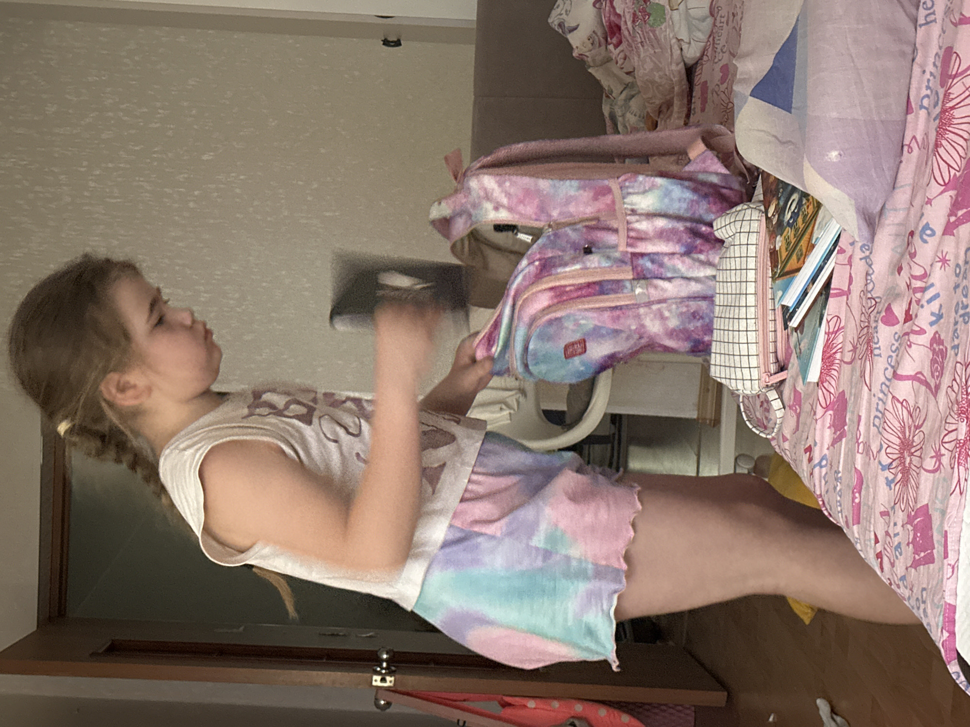 A young girl is depicted standing in what appears to be a bedroom in Chaikovsky, Russia. She has light brown hair, pulled back into a braid or pigtail, and is wearing a white sleeveless top paired with tie-dye shorts in shades of pink, purple, and blue. Her gaze is directed towards a colorful pink and blue tie-dye backpack, which is positioned on a bed to her right. The backpack features multiple zippered compartments and has a visible 'Skip' logo on one of its pockets. Attached to the backpack is a white pouch or pencil case with a black grid pattern, from which several books or notebooks can be seen protruding.
The bed is covered with patterned pink and white bedding, including text such as 'Princess' and various floral motifs. A separate light blue and white patterned blanket or pillow is also visible on the bed. The room features light-colored walls and a wooden floor. The girl's hands are actively engaged with the backpack, suggesting she might be in the process of packing or unpacking its contents. Her expression is focused, indicating concentration on the task at hand. The time of day and weather are not discernible from the image.