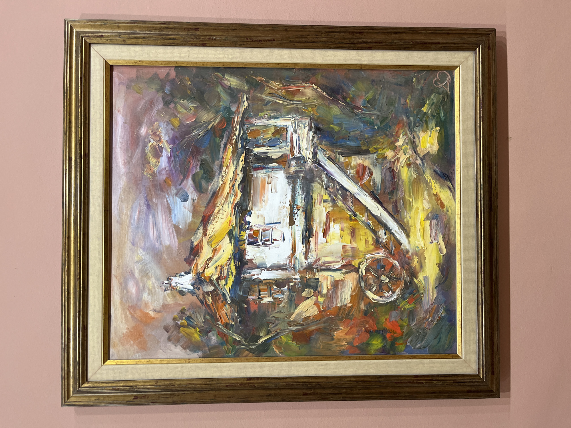 An oil painting, rendered in an impasto and impressionistic style, is depicted hanging on a plain, light pink wall. The artwork is framed within an ornate, multi-layered golden frame that includes an inner cream-colored linen liner.
The painting itself features a vibrant and somewhat abstract depiction of what appears to be an industrial structure, possibly a mining headframe or a similar machinery complex. The central elements of the structure are painted with thick, bright strokes of white, yellow, and orange, suggesting illumination or the primary focus. Recognizable components include a tall, tower-like section in the center, a sloped ramp or conveyor belt extending to the right, and a distinct wheel mechanism on the right side. The surrounding areas and background of the painting are rendered in darker, more abstract brushstrokes of blues, greens, browns, and hints of red and purple, creating a dynamic contrast with the bright central subject.
In the top right corner of the painting, a faint, stylized mark or signature, possibly resembling a heart or intertwined letters, is visible. No specific activity is shown within the painting; it portrays a static industrial scene. The overall scene suggests an art display, possibly within a home or gallery. The provided location context is Plovdiv, Bulgaria.