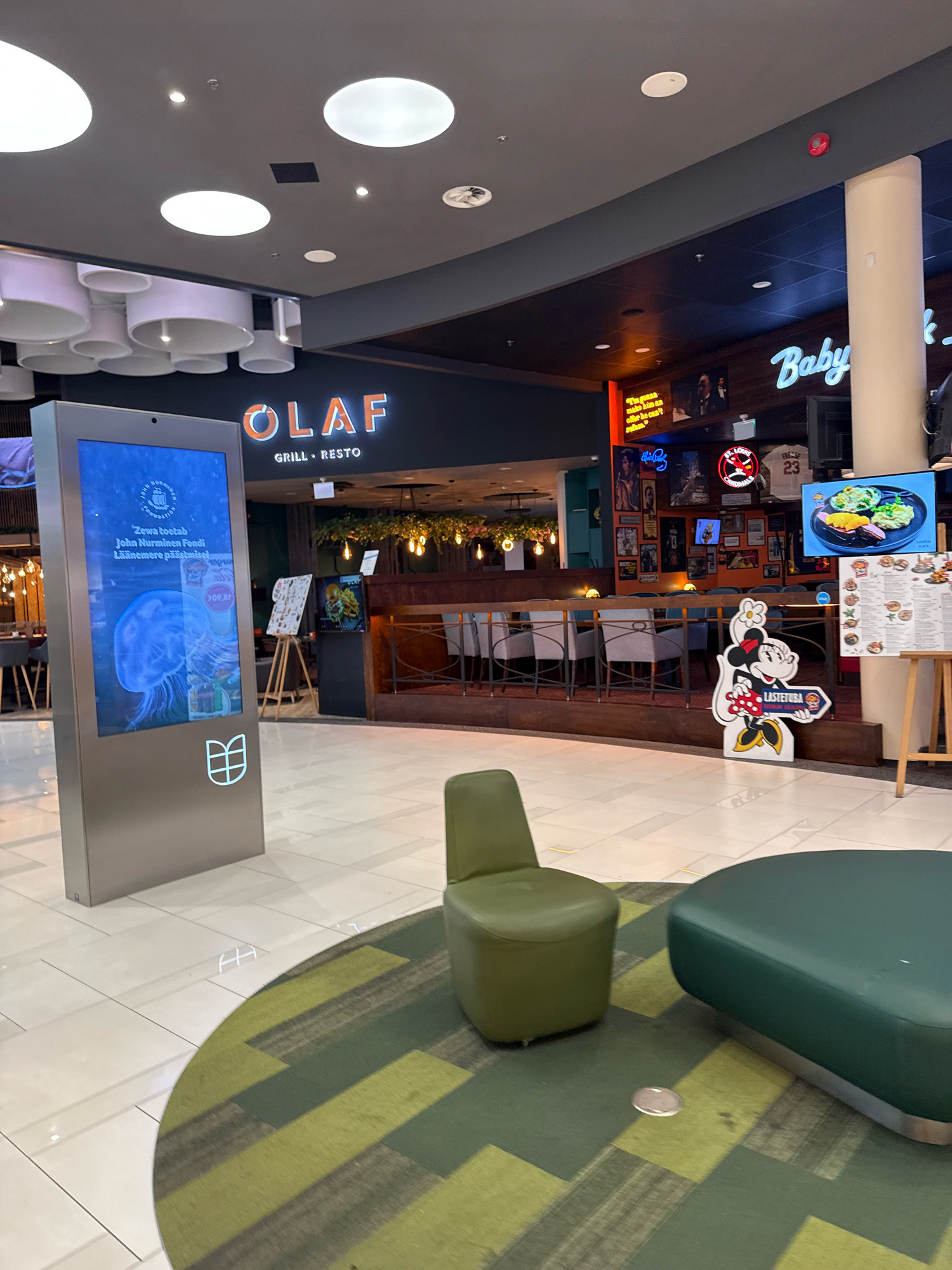 The image shows the interior of a modern shopping mall in Tallinn, Estonia. In the foreground, there is a large digital display screen with information about "John Nurminen Foundation" and a picture of a jellyfish. To the right, a green upholstered ottoman and a matching chair are placed on a circular rug with a green and brown geometric pattern. The background features the entrance to a restaurant named "OLAF GRILL•RETO," decorated with neon signs and posters. A large pillar and a column with exposed pipes are visible on the right side of the restaurant entrance. Various tables and chairs are set up for dining inside the restaurant. The mall floor is tiled with light-colored square tiles.