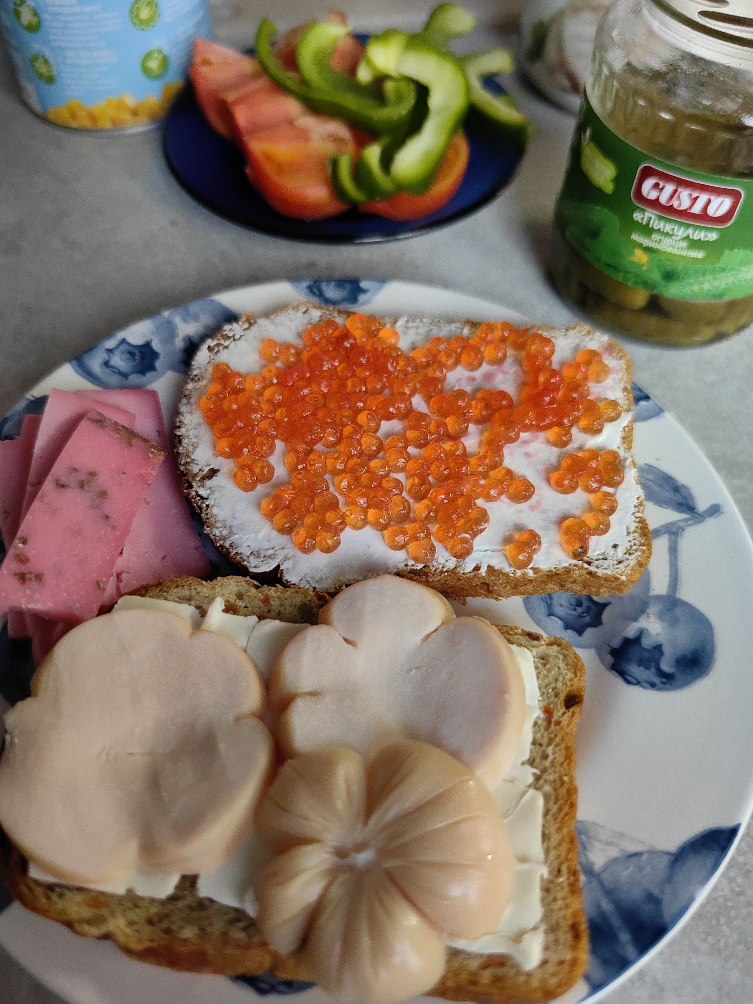 This is a close-up, eye-level, overhead shot of a plate of food. The plate is white with a blue floral pattern. On the plate, there is a slice of brown bread topped with butter and red caviar. Next to it, another slice of brown bread is topped with butter and some sliced pink cheese. In the foreground, there are two more slices of brown bread topped with butter and sliced meat that is shaped like flowers. In the background, a blue plate holds sliced tomatoes and green bell peppers. A jar of pickles with a green label that says "Custo" and "Пикули" is also visible in the background. The image appears to be taken indoors, possibly in a kitchen or dining area. There is no indication of the location being in the city of Fanipal, Belarus.
