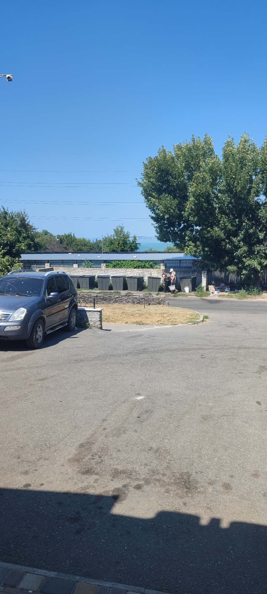 In Svitlovodsk, Ukraine, under a clear blue sky, a dark blue SUV is parked on a paved area. Adjacent to the SUV, a row of dark rectangular bins stands behind a low fence constructed of interwoven branches. Two individuals are actively engaged near these bins. One person, wearing a light-colored cap and a patterned shirt, appears to be assisting or interacting with another person who is facing away from the camera, wearing a dark shirt and pants. A large, leafy tree dominates the right side of the scene, casting a shadow on the pavement in the foreground. In the background, a line of solar panels is visible, and beyond that, a body of water and more trees can be seen. A security camera is mounted on a pole on the left side of the image, facing towards the sky.