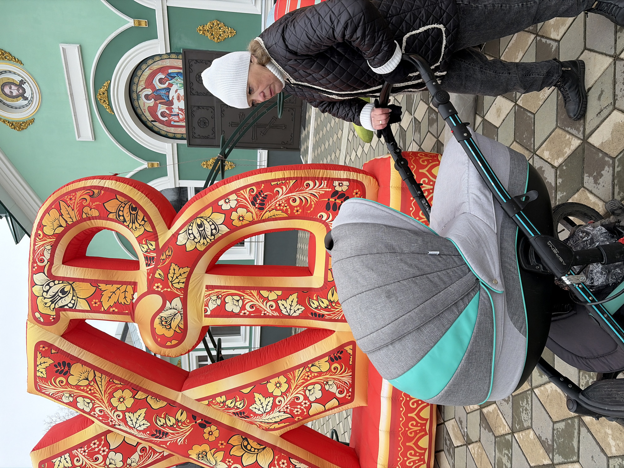 A woman with a stroller is standing in front of the Saint George temple in Chaikovsky, Russia. The woman is wearing a black jacket, dark jeans, and black boots. She is looking down at the stroller. The stroller is gray and black, and has a green trim. The temple is green and white with gold accents. There is a large red and gold sign in front of the temple. The ground is paved with gray and brown patterned bricks.