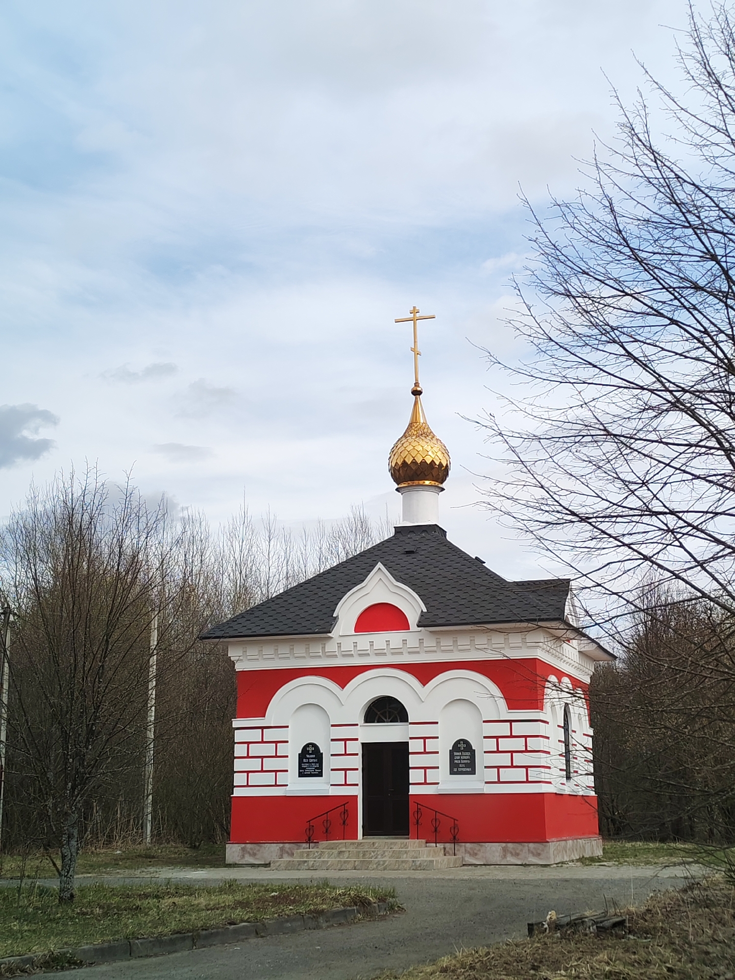 The image shows a small red and white Orthodox church or chapel located in Unknown, Unknown. The building has a distinctive design with a red base, white brickwork accents, arched windows, and a black roof. A golden dome with a cross on top sits above the roof. The chapel is surrounded by bare trees and vegetation. A paved pathway leads to the entrance of the chapel. The sky is overcast.