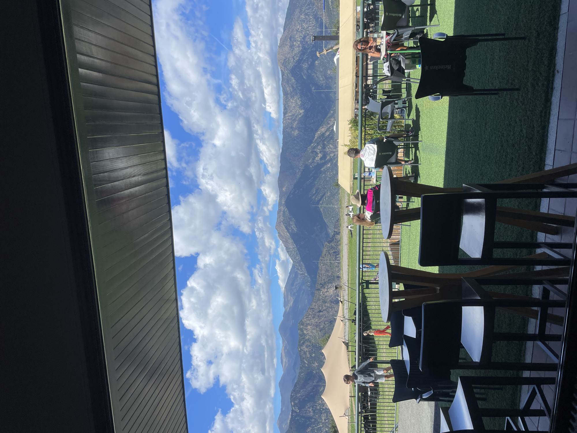 The image depicts an outdoor seating area of a restaurant or cafe, likely located in Juberri, Andorra, with a backdrop of imposing mountains and a cloudy sky. In the foreground, several dark, modern chairs and tall, wooden tables are arranged on a green, artificial turf surface. Some of these chairs have "Heineken" branding visible.
Further into the scene, a railing separates the seating area from a larger open space. Beyond the railing, a few people are seated at tables and walking around. A notable structure is a large, light-colored tent situated in the middle ground. In the distance, a range of rugged mountains forms the horizon, with their peaks catching the sunlight. The sky is a mix of blue and white, with scattered clouds.
A young boy in a grey hoodie and shorts is seen walking away from the camera towards the tent. Another individual, a woman with dark hair, is seated at a table on the right, looking at her phone. The overall atmosphere appears relaxed and casual, suggesting a daytime leisure setting. The architectural elements in the foreground, such as the overhang and its ribbed underside, indicate the structure is a covered patio or balcony of a building.